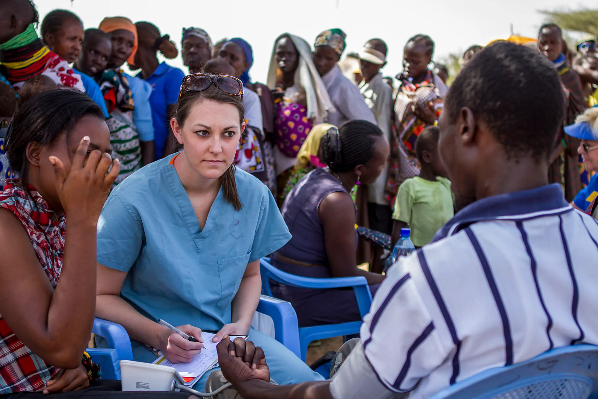Healthcare worker in blue scrubs listens intently and takes notes while holding the hand of a seated patient outdoors with a group of people nearby.