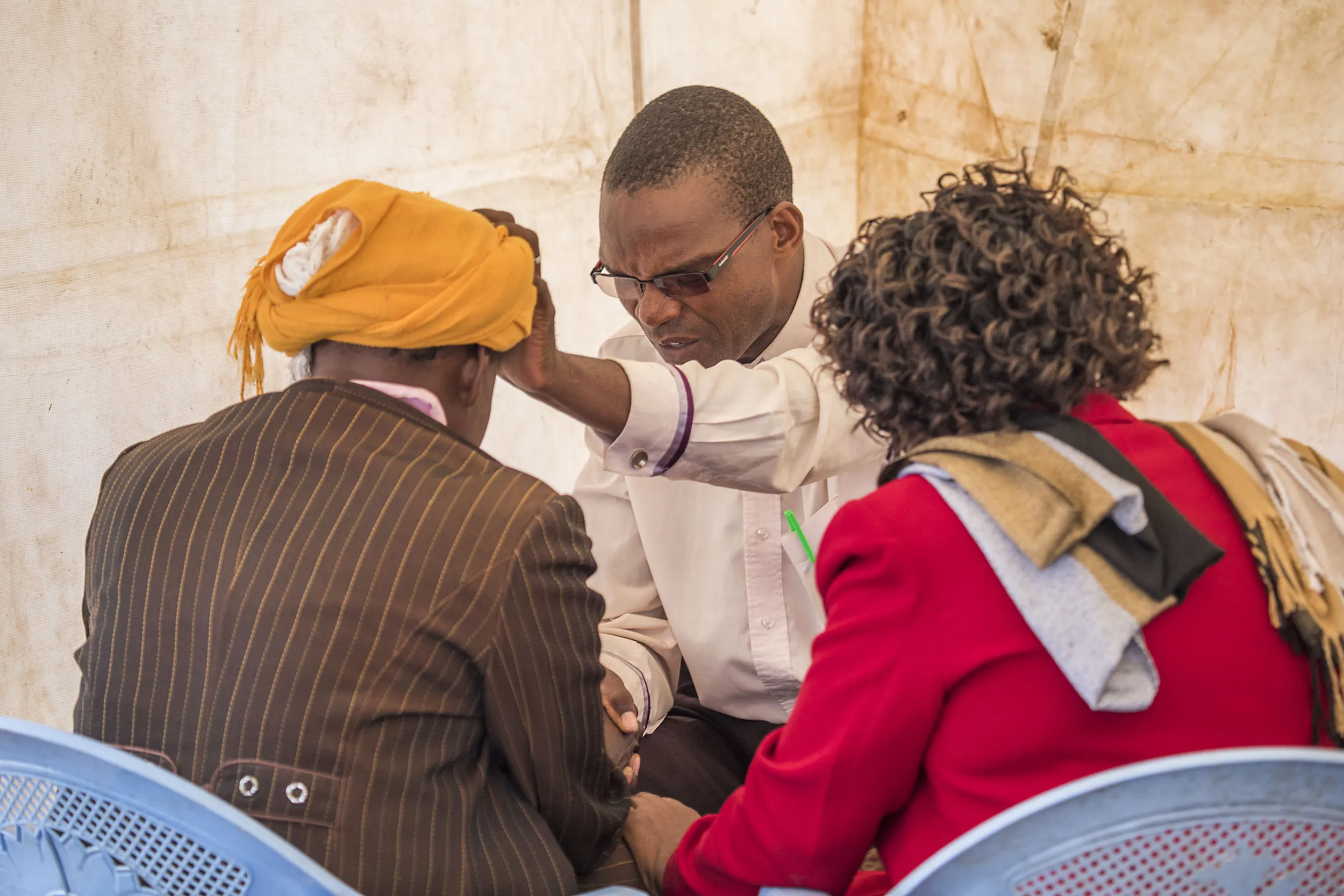 Man with glasses holding another man's head in a blessing or prayer gesture, with a woman seated beside them.