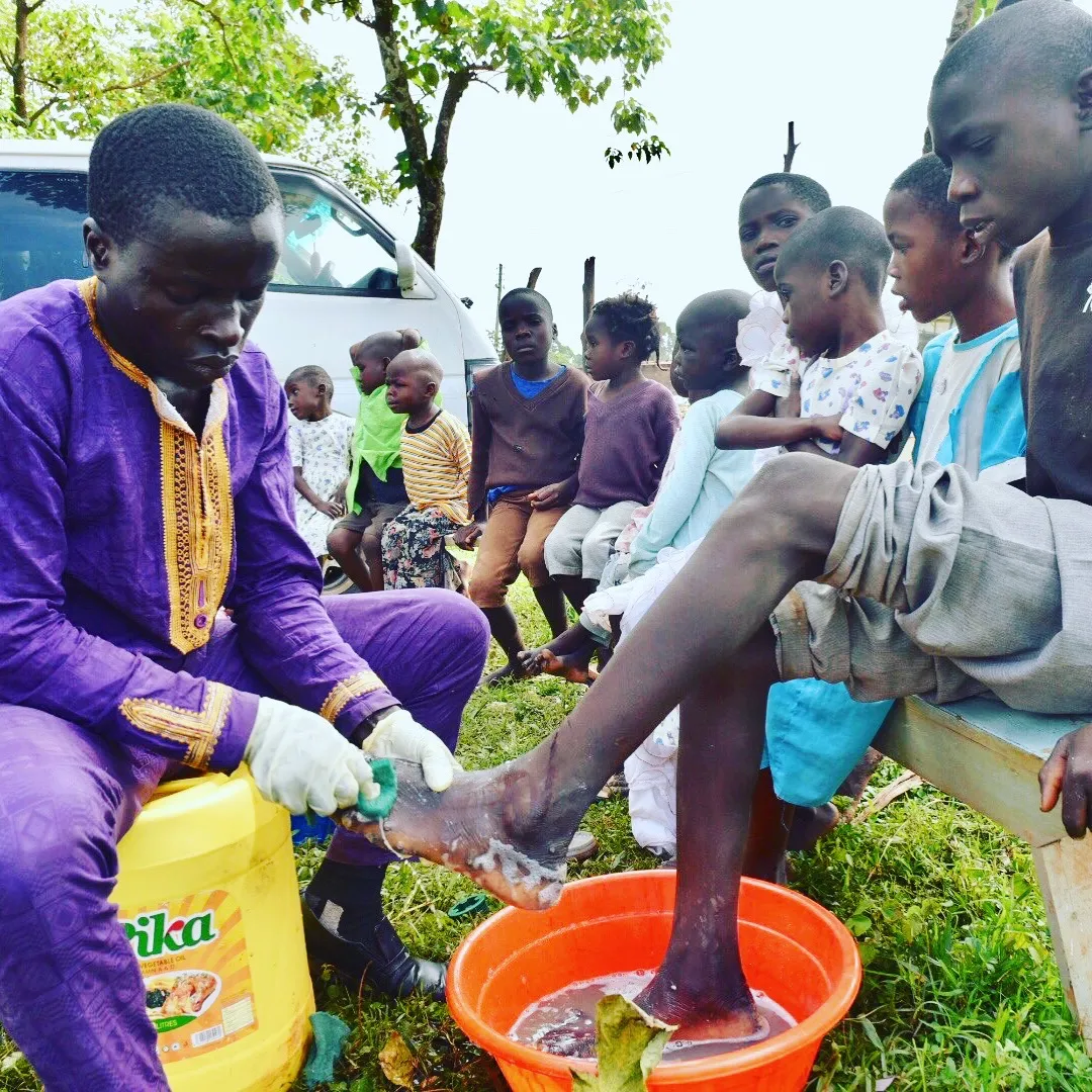 A man in a purple traditional outfit washes the foot of another person seated on a bench, while several children watch outdoors.