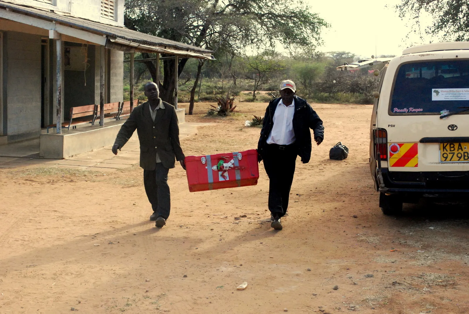 Two men carrying a large red container on a dirt path near a building and a white van.