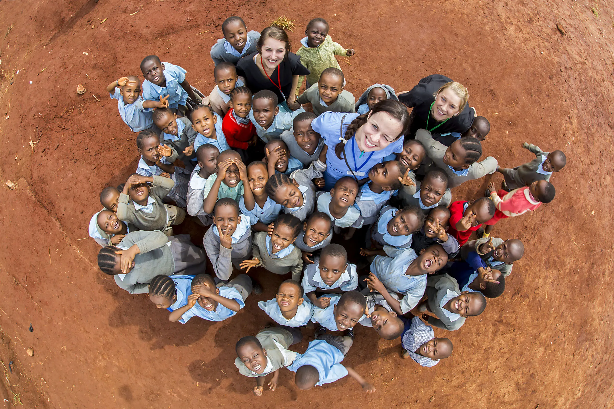 Group of smiling children and three adults looking up, standing close together on red dirt ground.