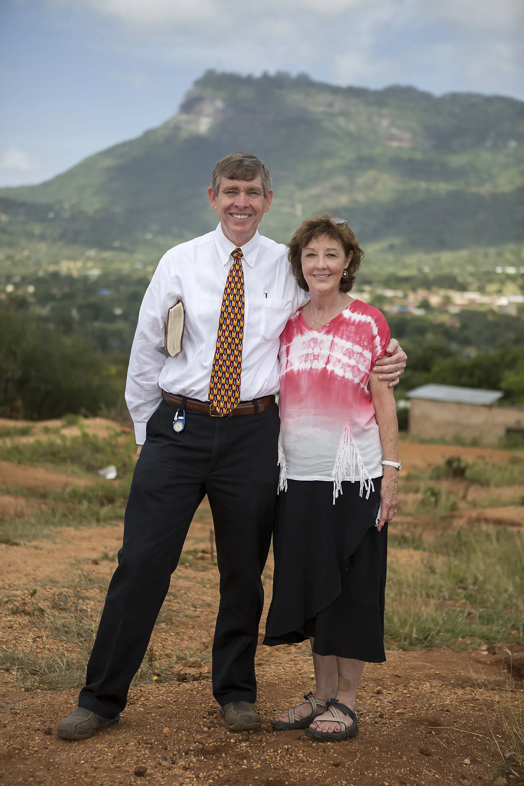 Smiling man and woman stand outdoors on a dirt path, with the man holding a book and a mountain in the background.