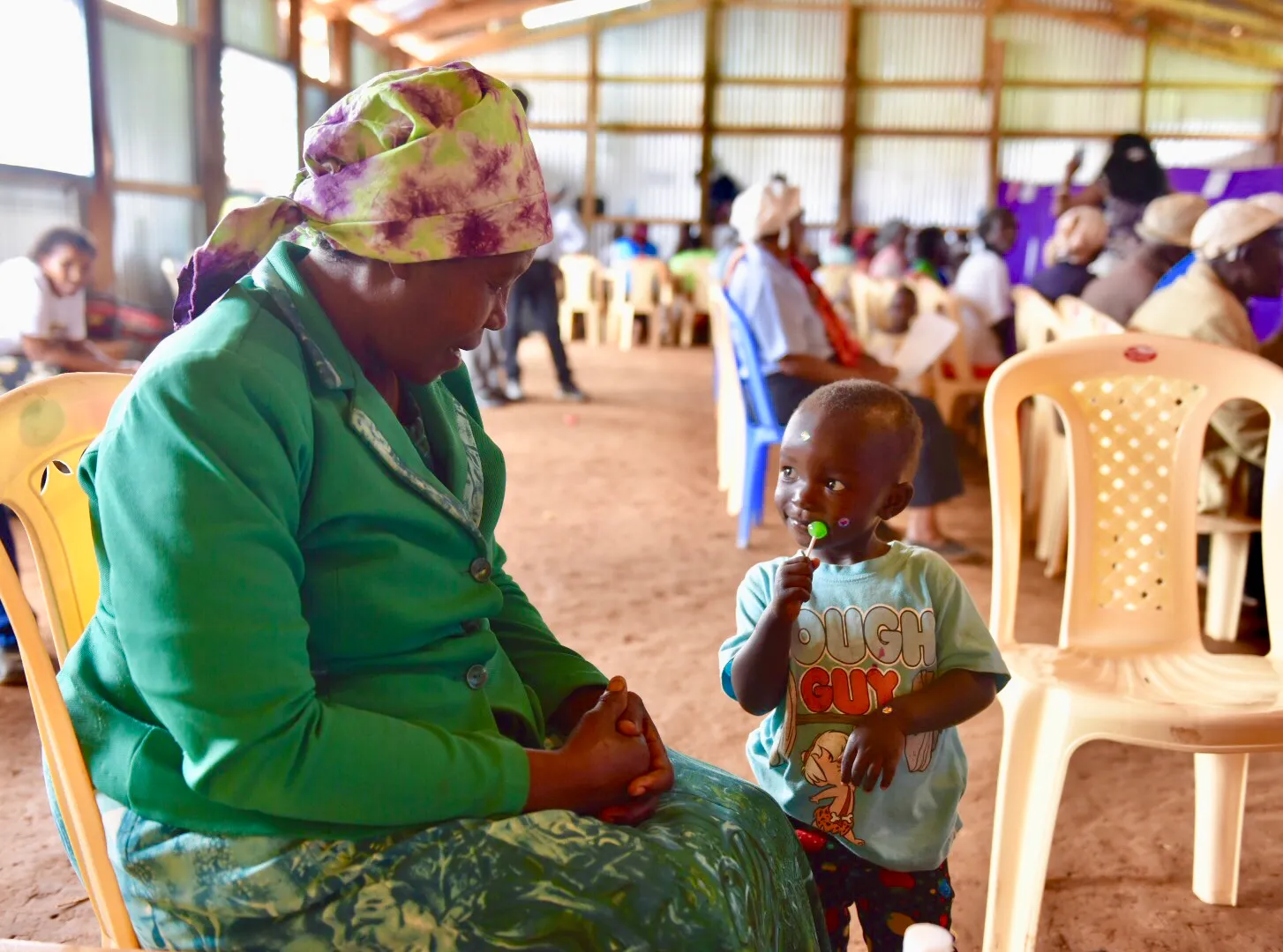 Woman in green jacket and headscarf sitting on chair, smiling at a young boy holding a green lollipop in a crowded indoor setting.
