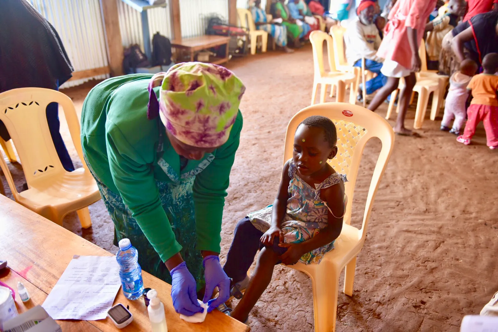 Healthcare worker wearing gloves cleaning a young girl's foot while she sits on a plastic chair indoors.