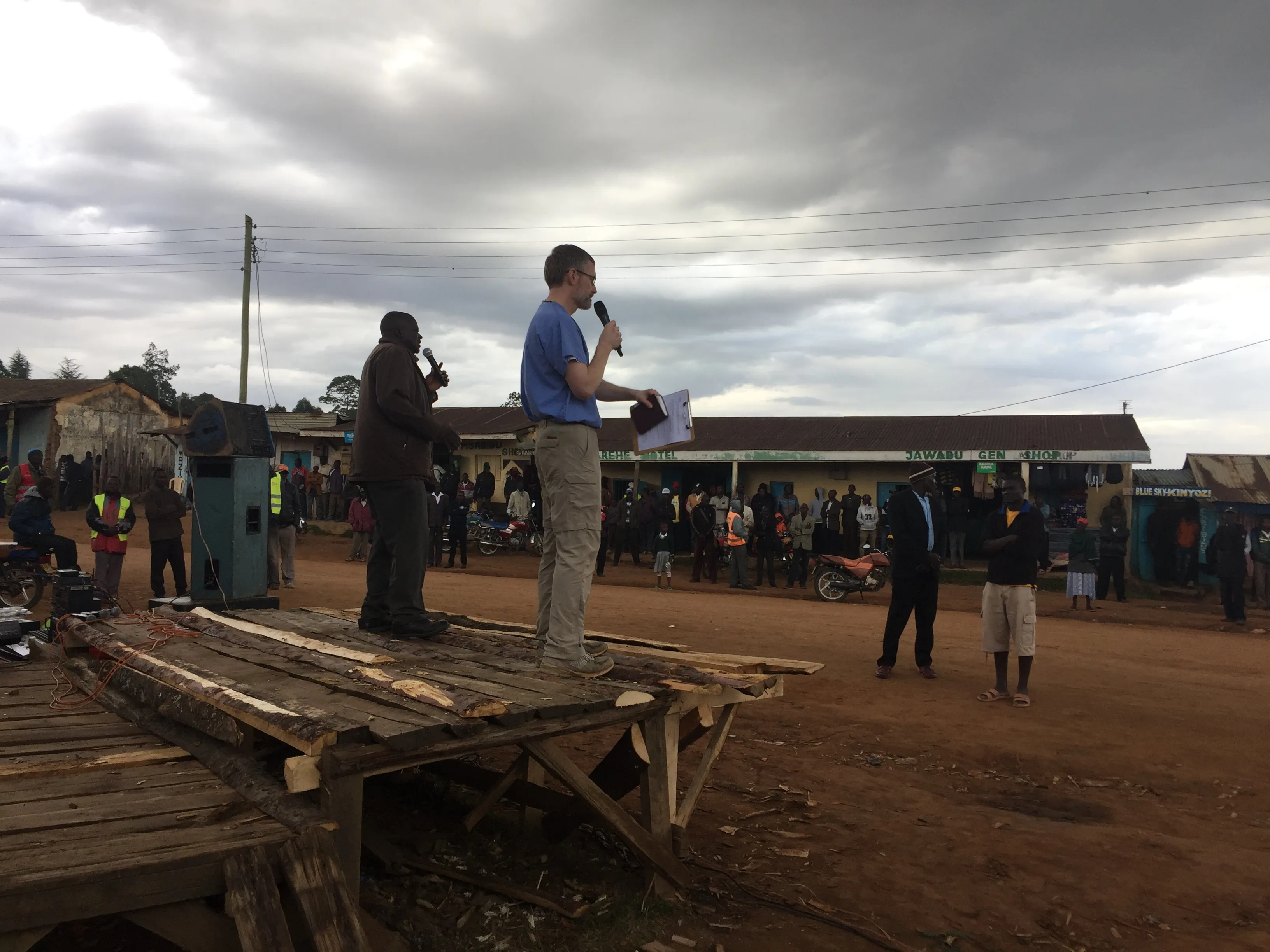Two men with microphones standing on a wooden platform addressing a crowd on a dirt road in a village with shops and motorcycles under a cloudy sky.