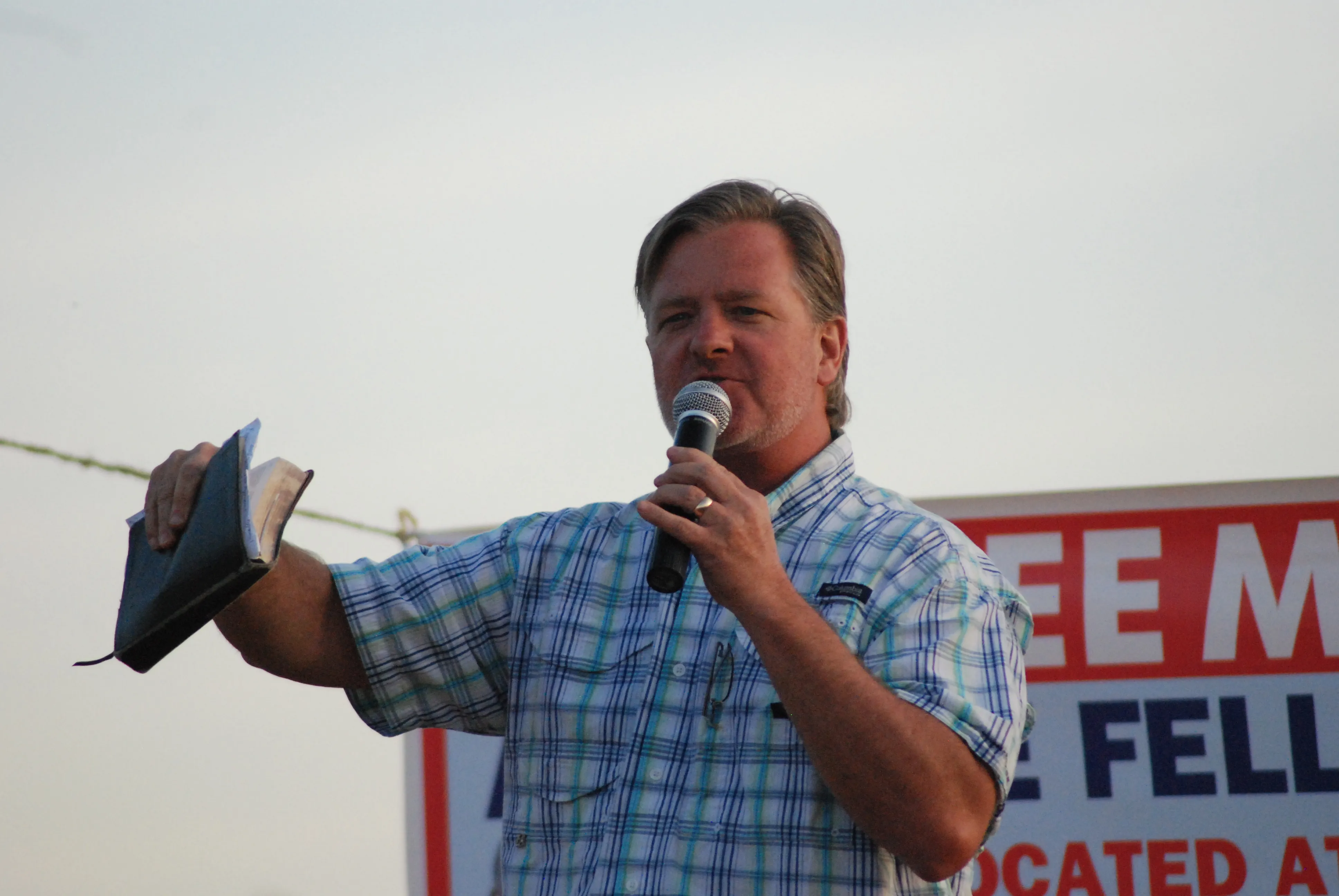 Man in plaid shirt speaking into a microphone and holding an open book outdoors.