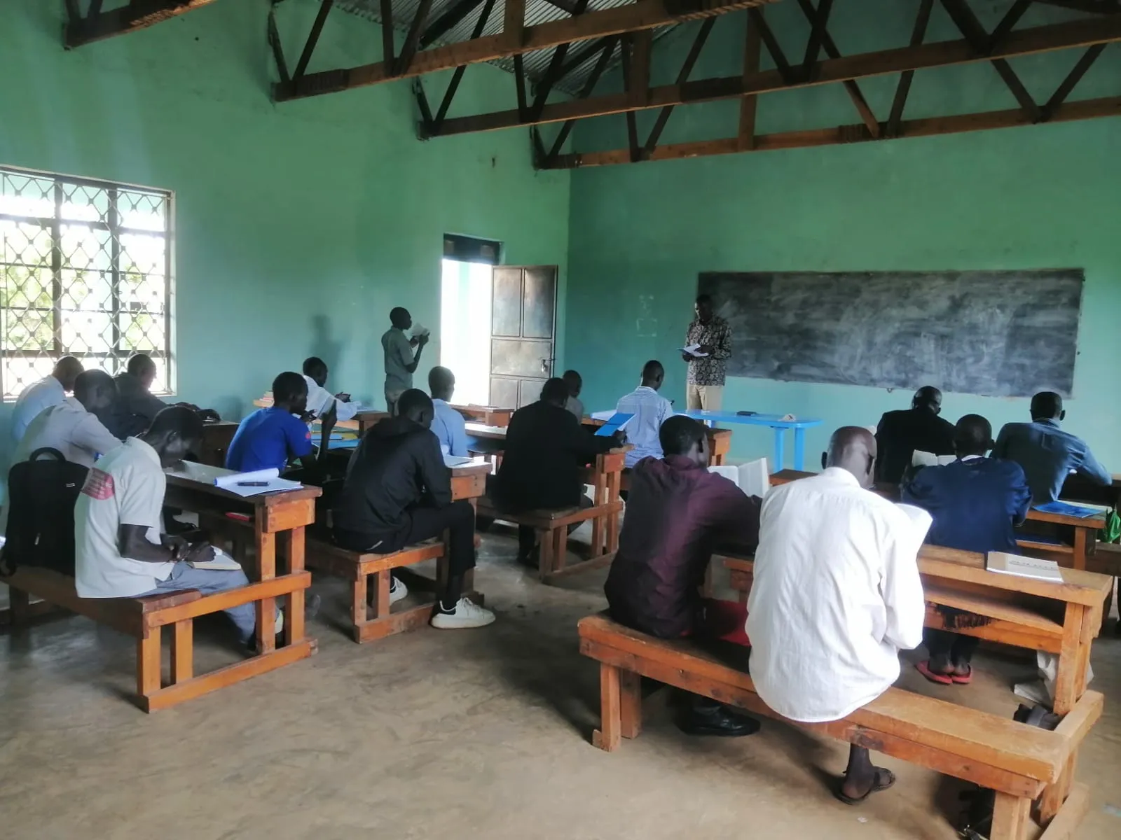 Classroom with adults seated on wooden benches facing a teacher standing near a chalkboard.