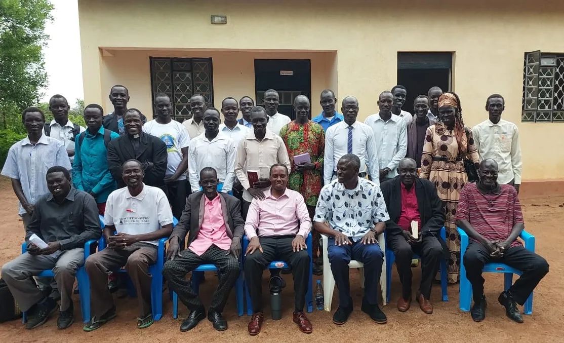 Group of men and one woman posing outdoors in front of a beige building, some seated on blue plastic chairs, others standing behind.