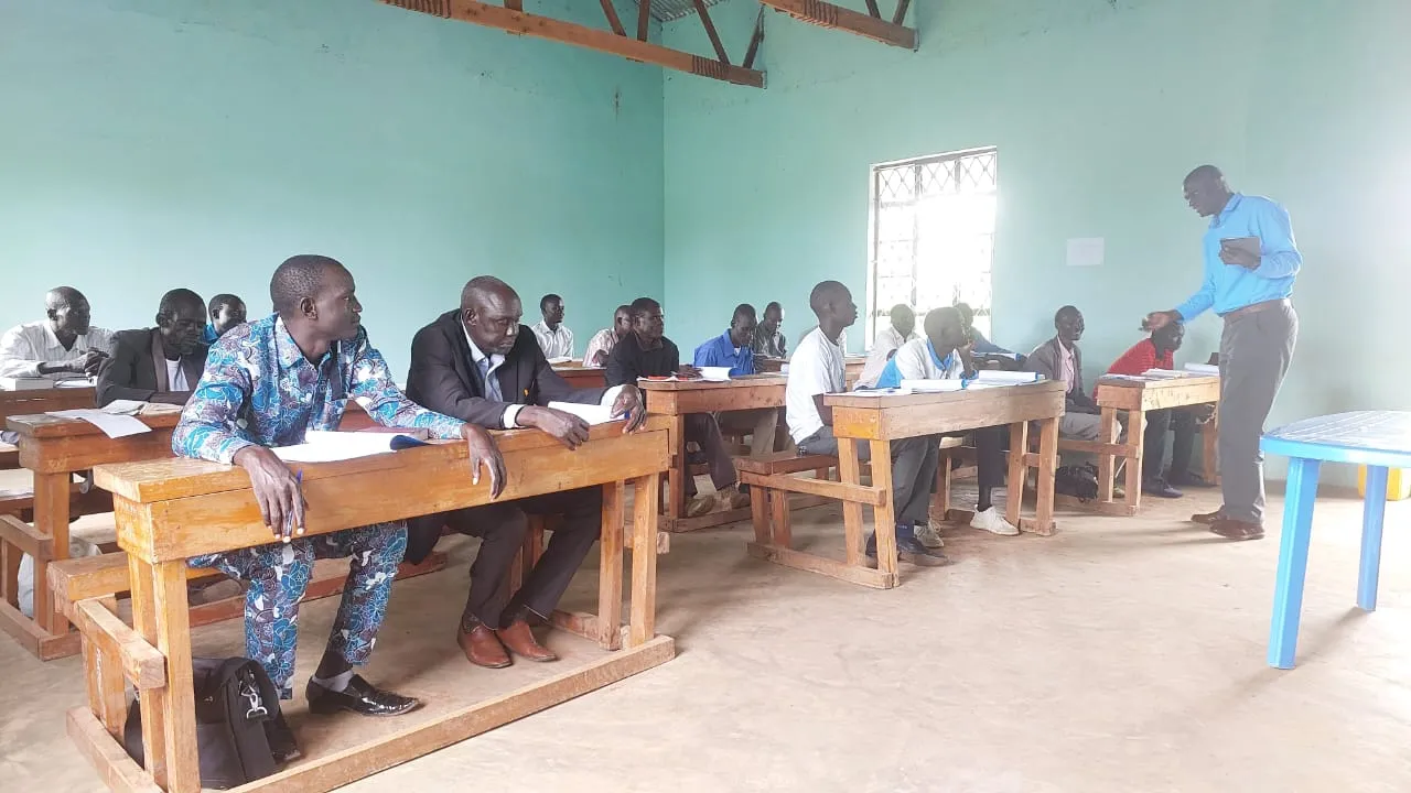 Adult men seated at wooden desks in a classroom listening to a standing instructor near a window.