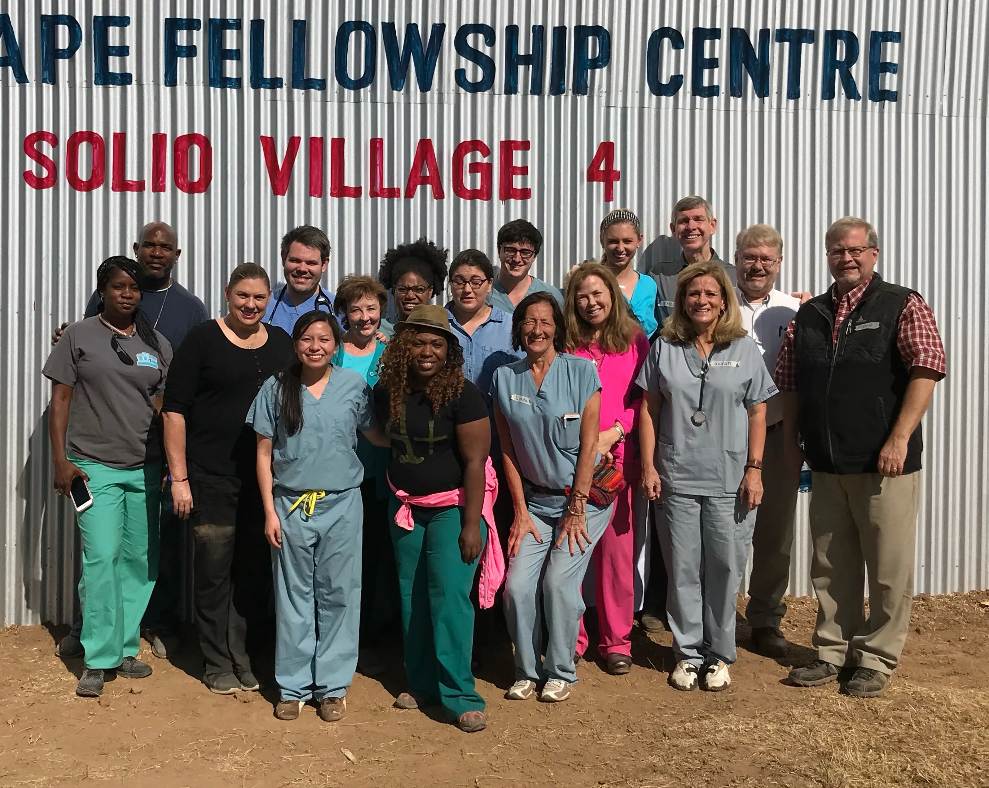 Group of diverse healthcare professionals and volunteers standing in front of a corrugated metal building with the sign 'Cape Fellowship Centre Solio Village 4.'
