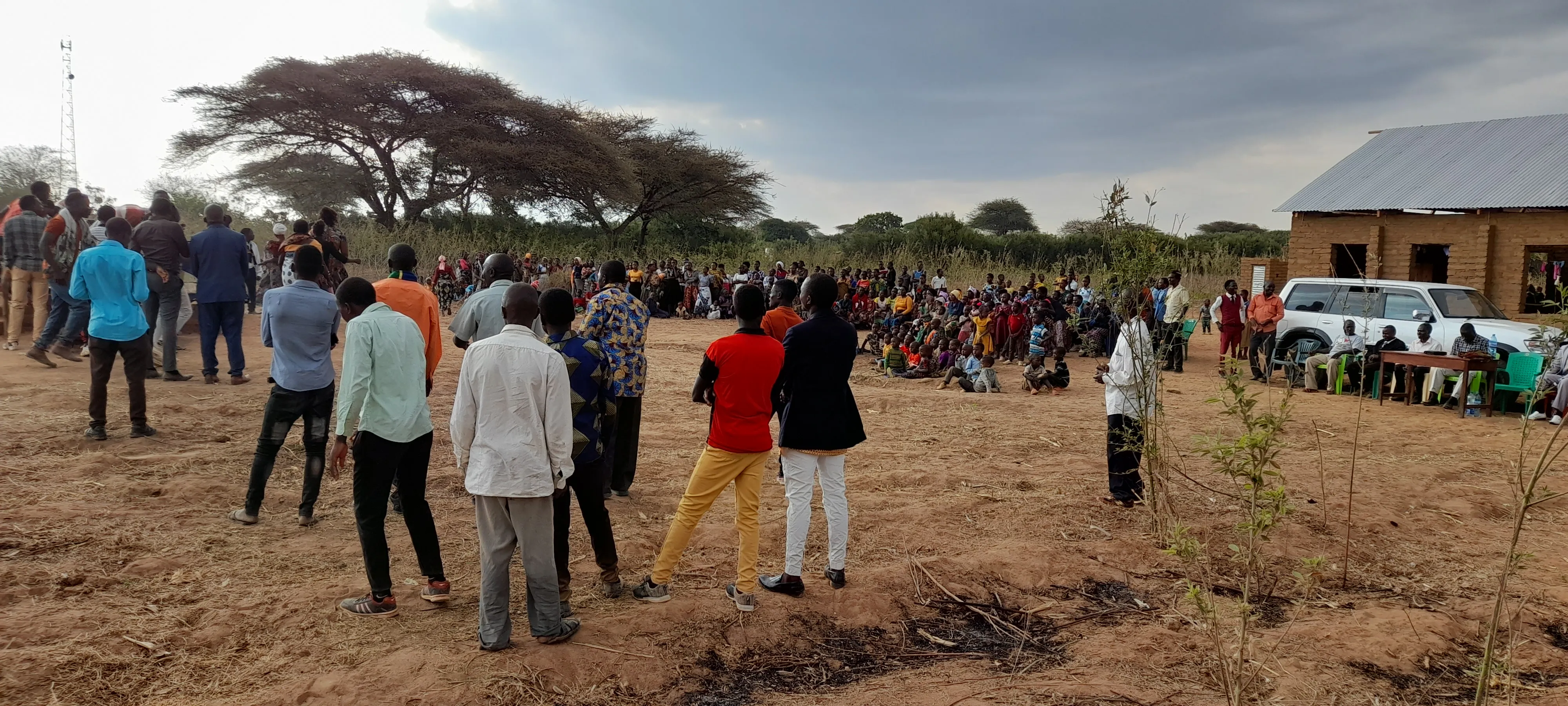 Large outdoor gathering of people, including men, women, and children, on dry ground near a building and a parked white vehicle under a cloudy sky.