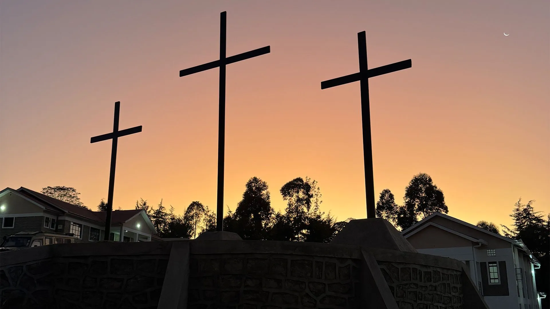 Three large crosses silhouetted against an orange sunset sky with trees and buildings in the background.