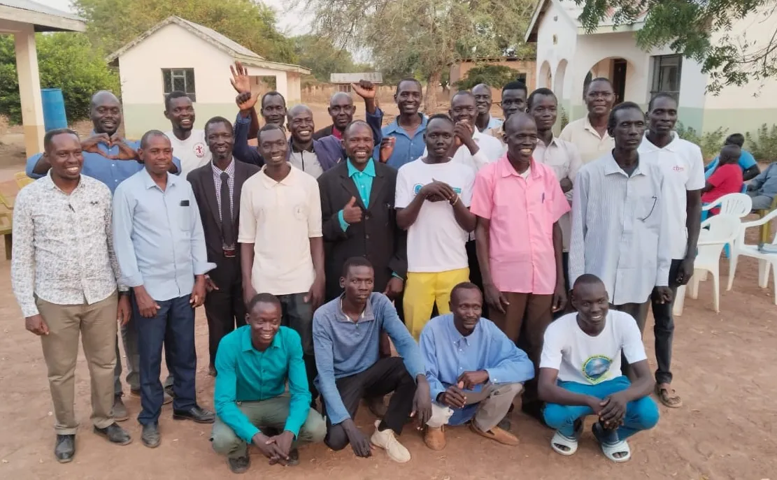 Group of men posing outdoors in front of buildings with trees in the background, some smiling and making hand gestures.
