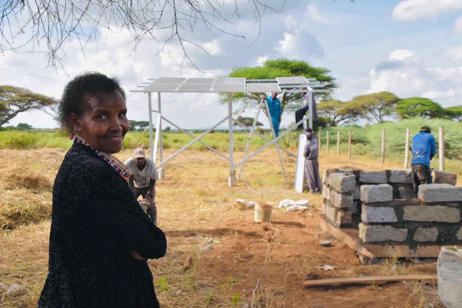 Woman smiling with arms crossed in foreground while four people work on assembling solar panels in a rural outdoor setting.