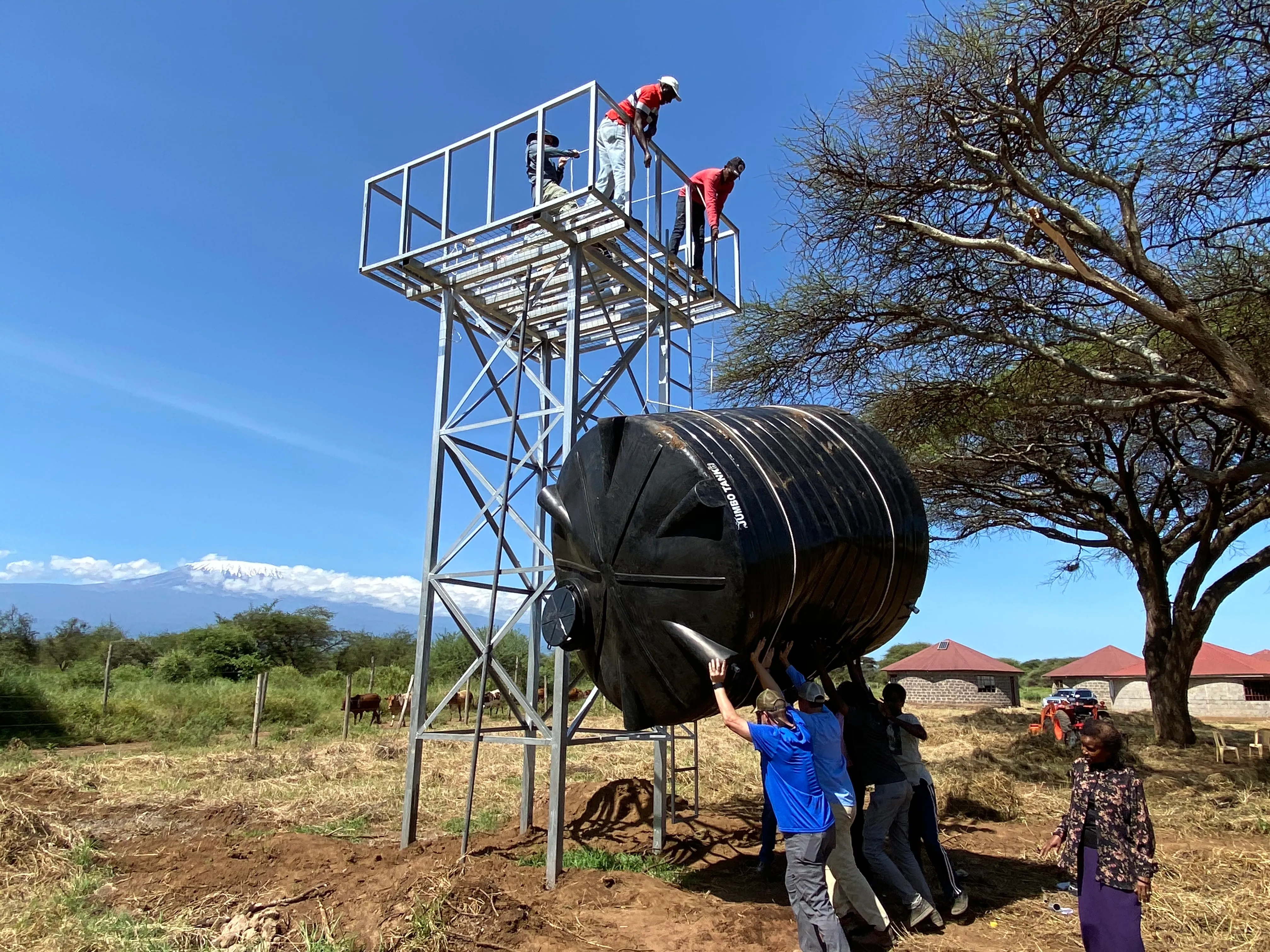 Group of people installing a large black water tank on a metal stand in a rural area with a clear sky and a mountain in the background.