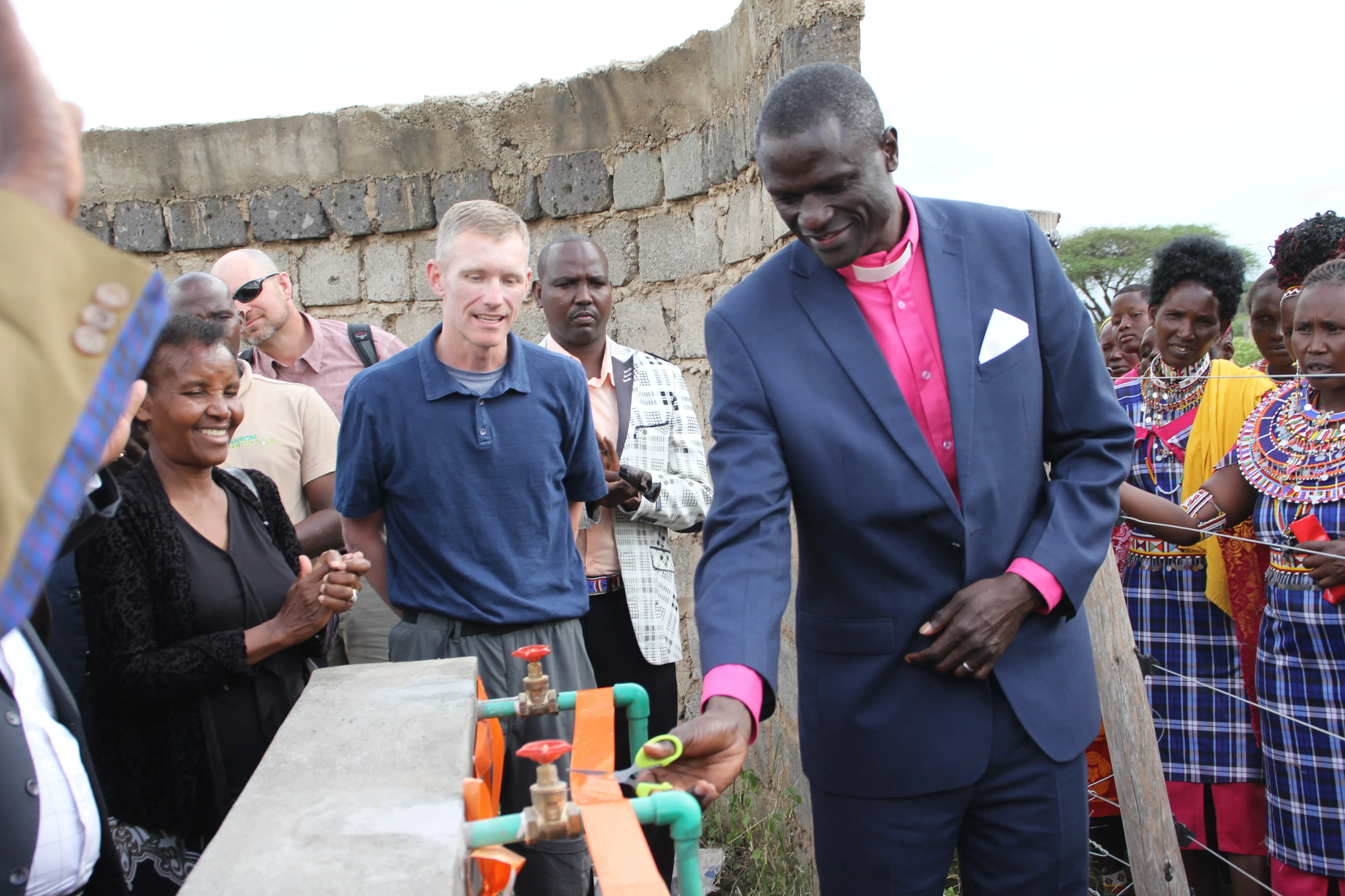 Man in a navy suit and pink clerical shirt cutting an orange ribbon at a water tap installation with a group of people watching.