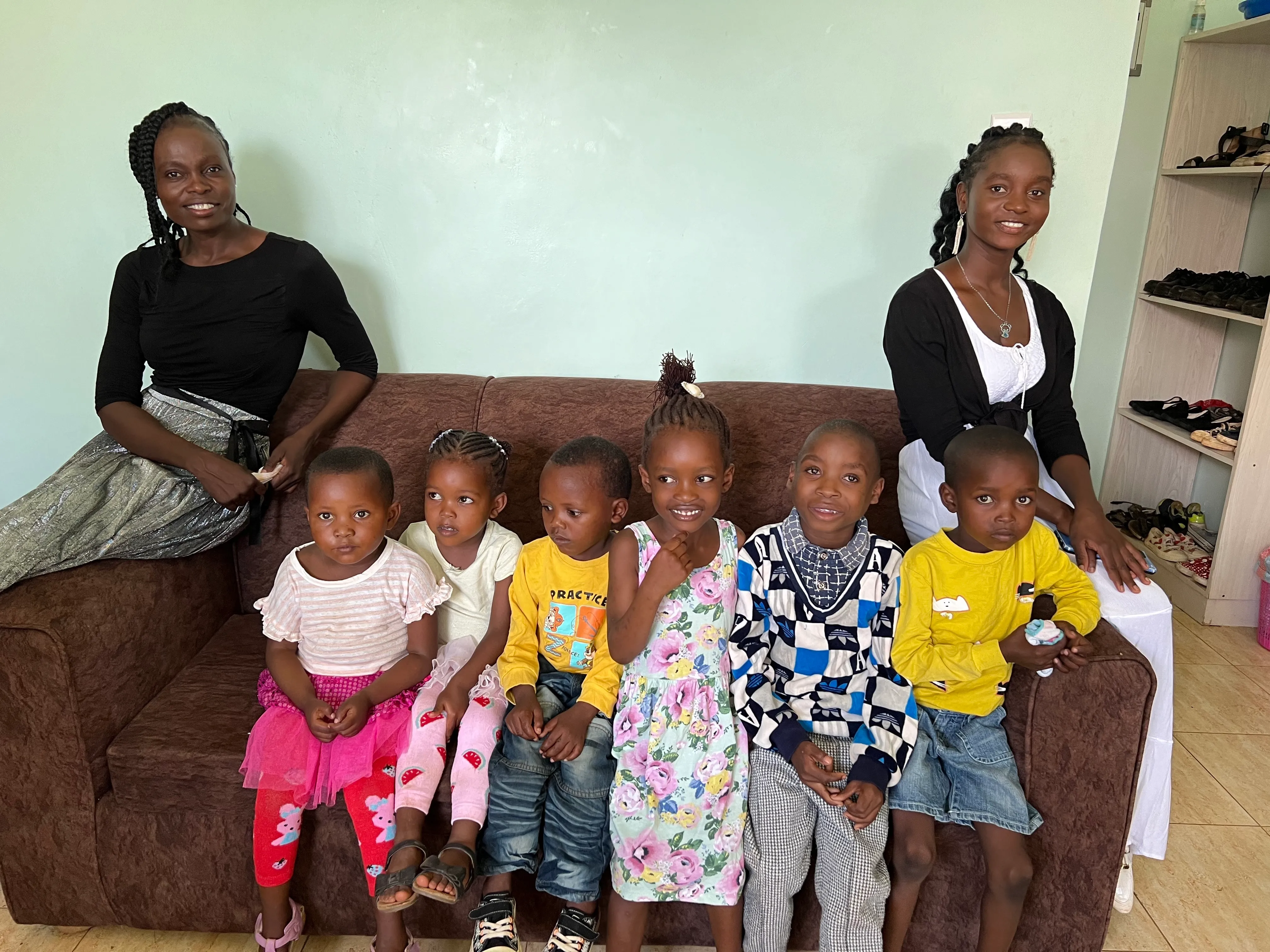 Two women sitting on a brown couch with six young children seated between them in a room with light green walls and a shoe rack visible in the background.