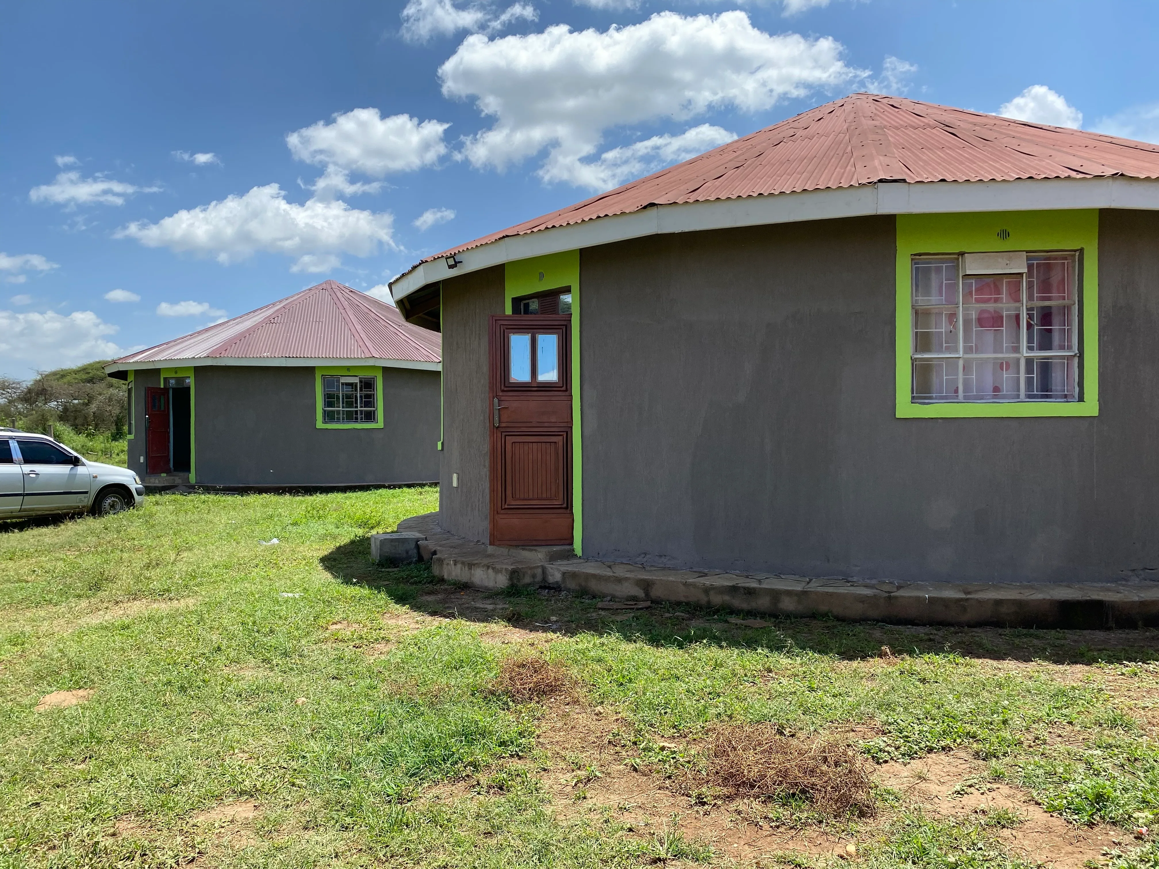 Two round houses with gray walls, green window frames, and red metal roofs under a blue sky with clouds on a grassy plot.