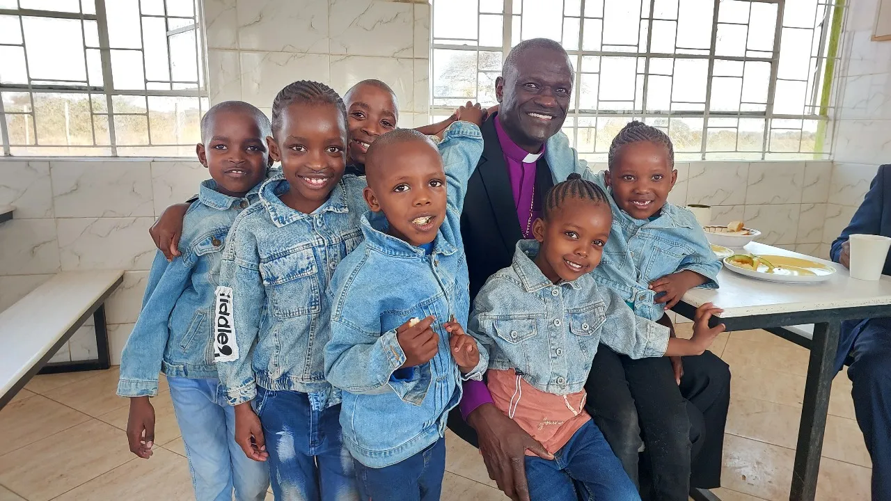 Smiling man in clerical collar surrounded by seven children wearing denim jackets inside a bright room.