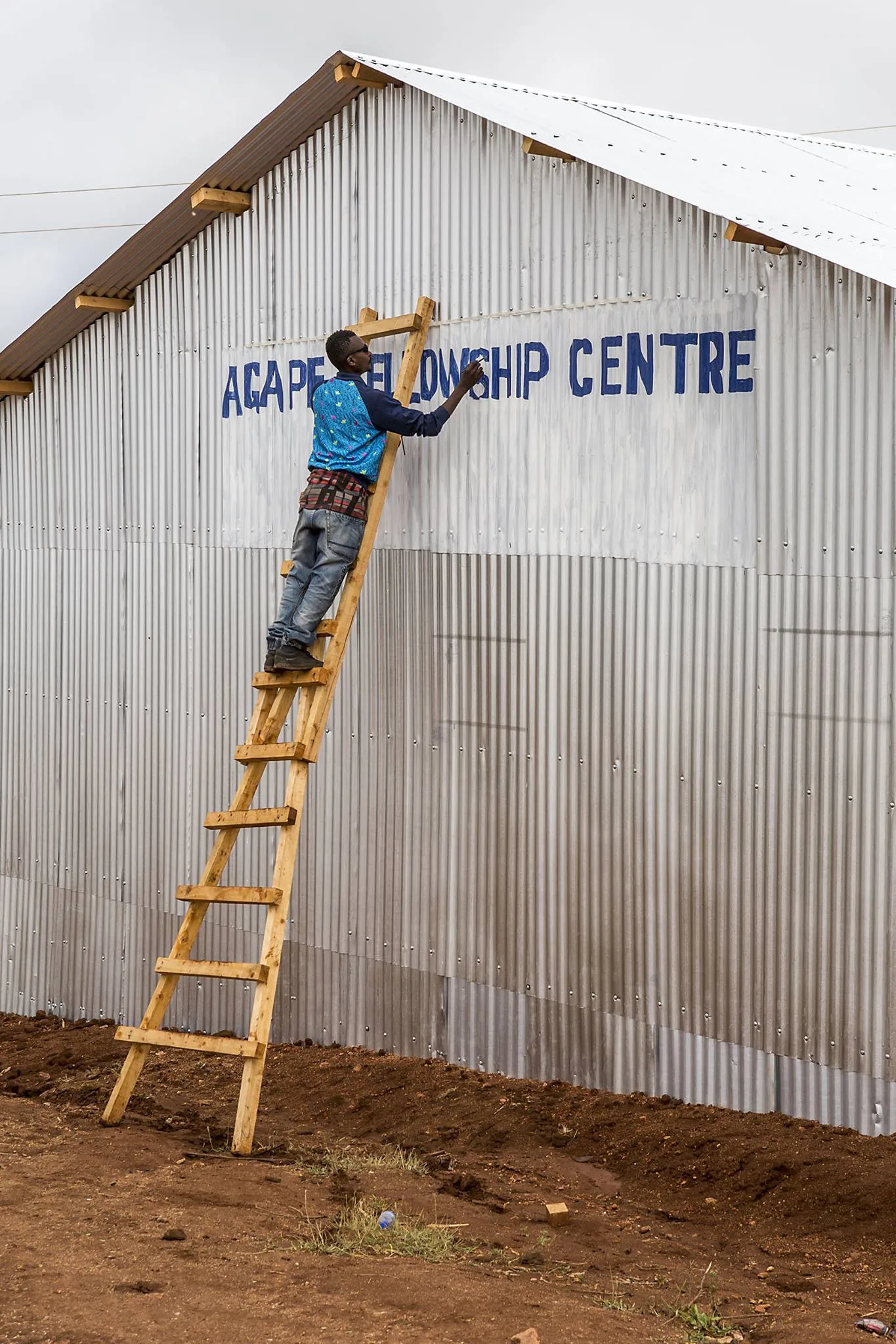 Man on a wooden ladder painting 'Agape Fellowship Centre' on a corrugated metal building.