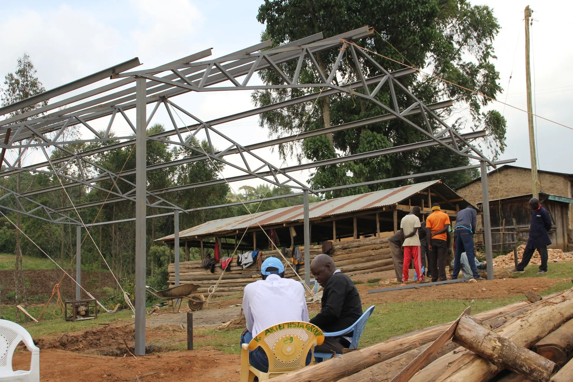 Two men sit on plastic chairs in front of a metal framework structure under construction with wooden logs and a group of workers nearby.