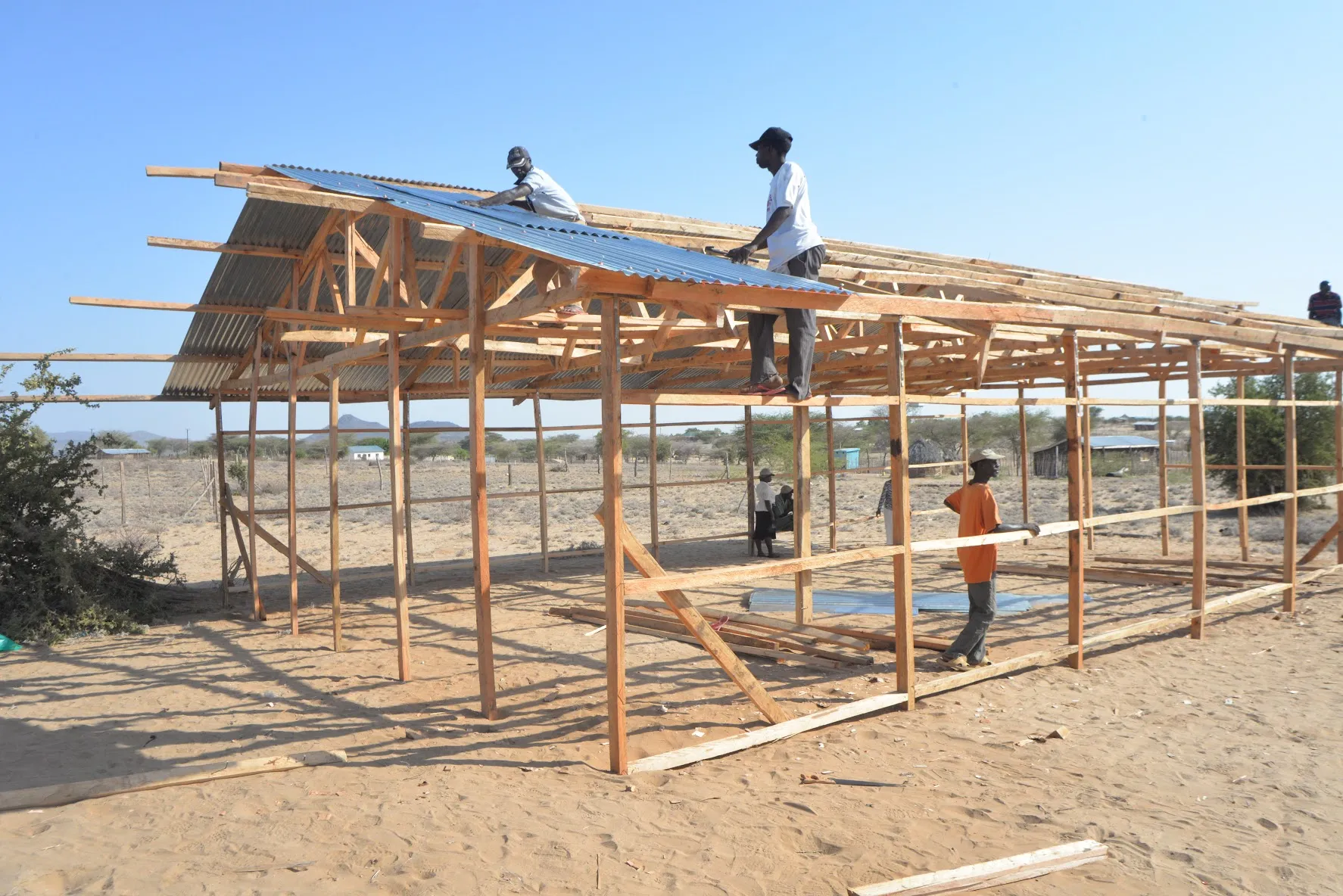 Men constructing a wooden frame building with corrugated metal roofing sheets in a dry, open area under a clear sky.