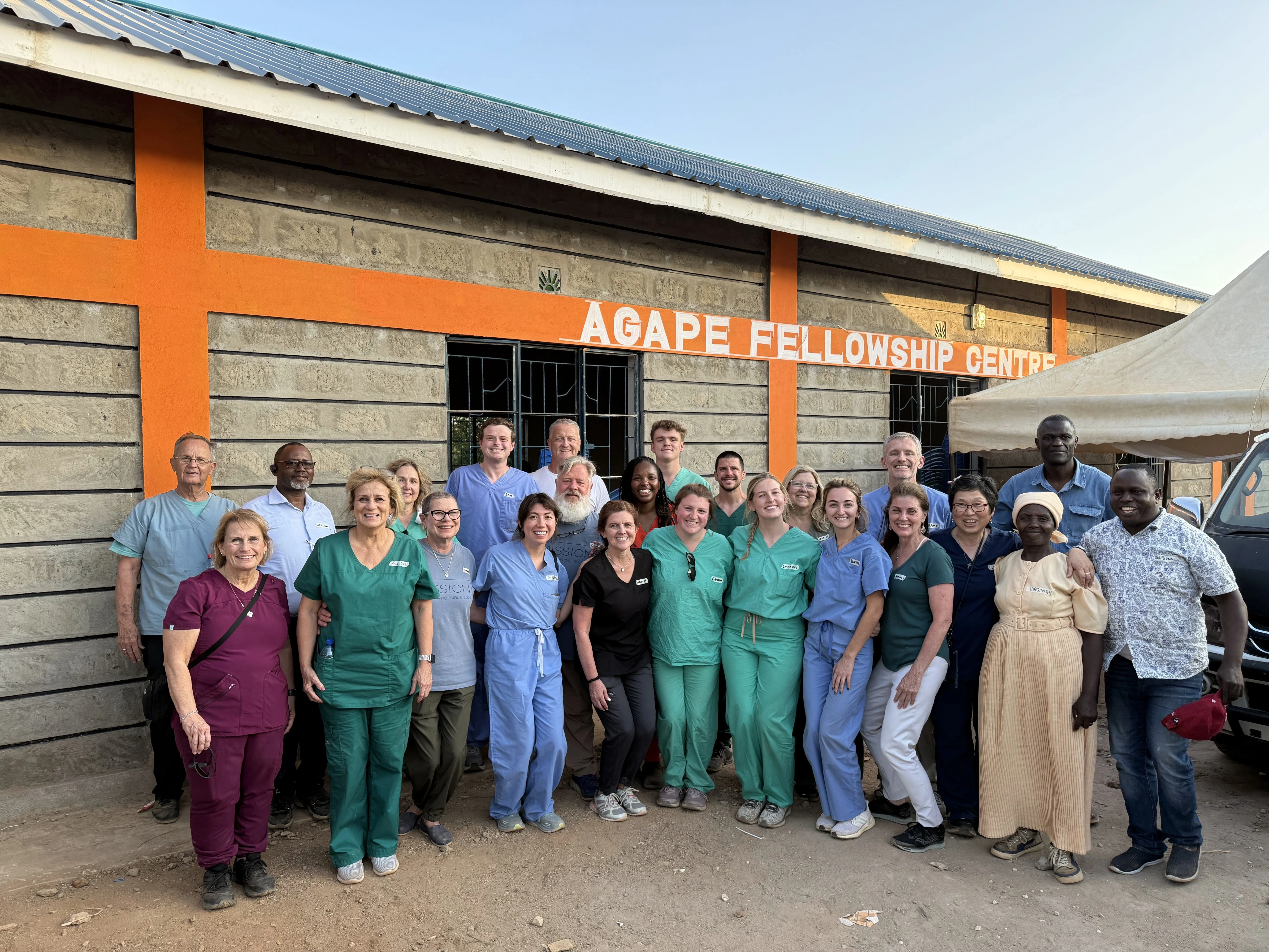 Group of healthcare workers and local community members posing outside Agape Fellowship Centre building with orange trim.