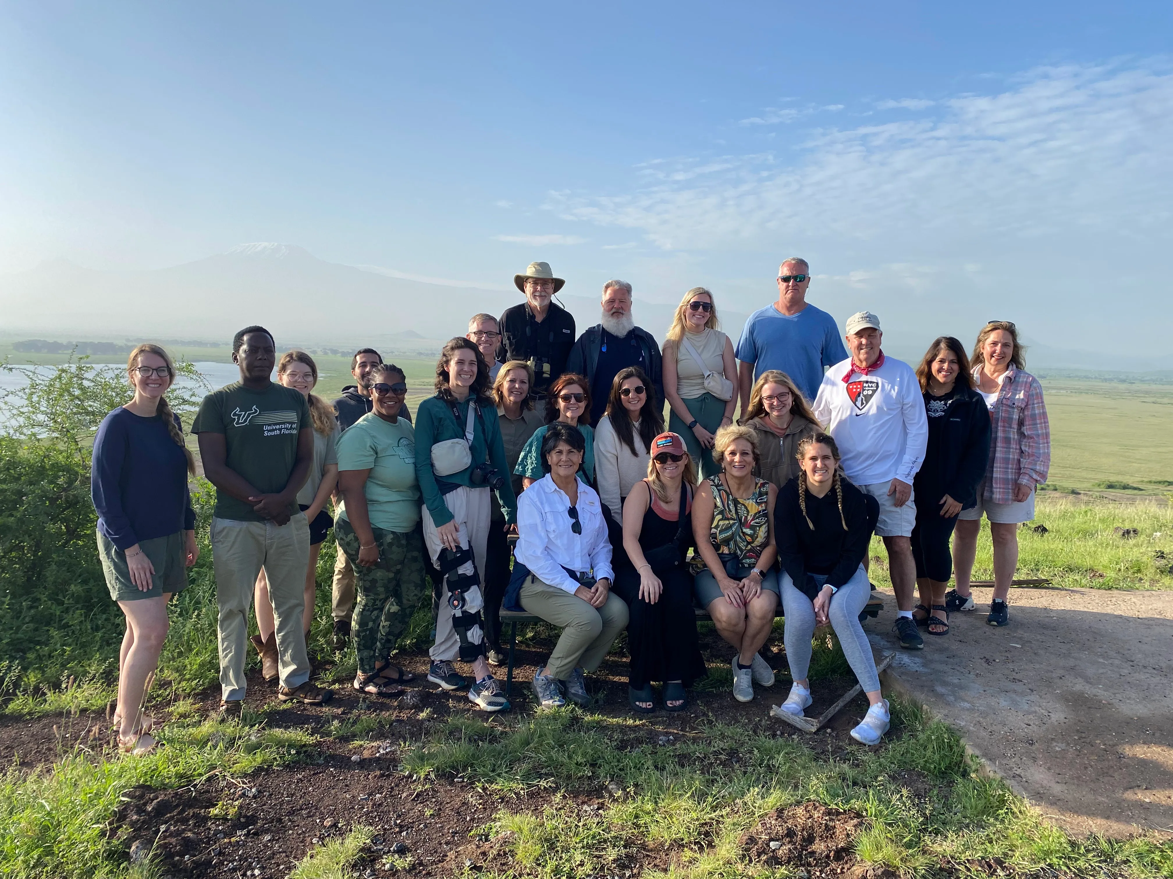 Large group of twenty people posing outdoors in front of a grassy plain with a mountain in the background under a clear blue sky.