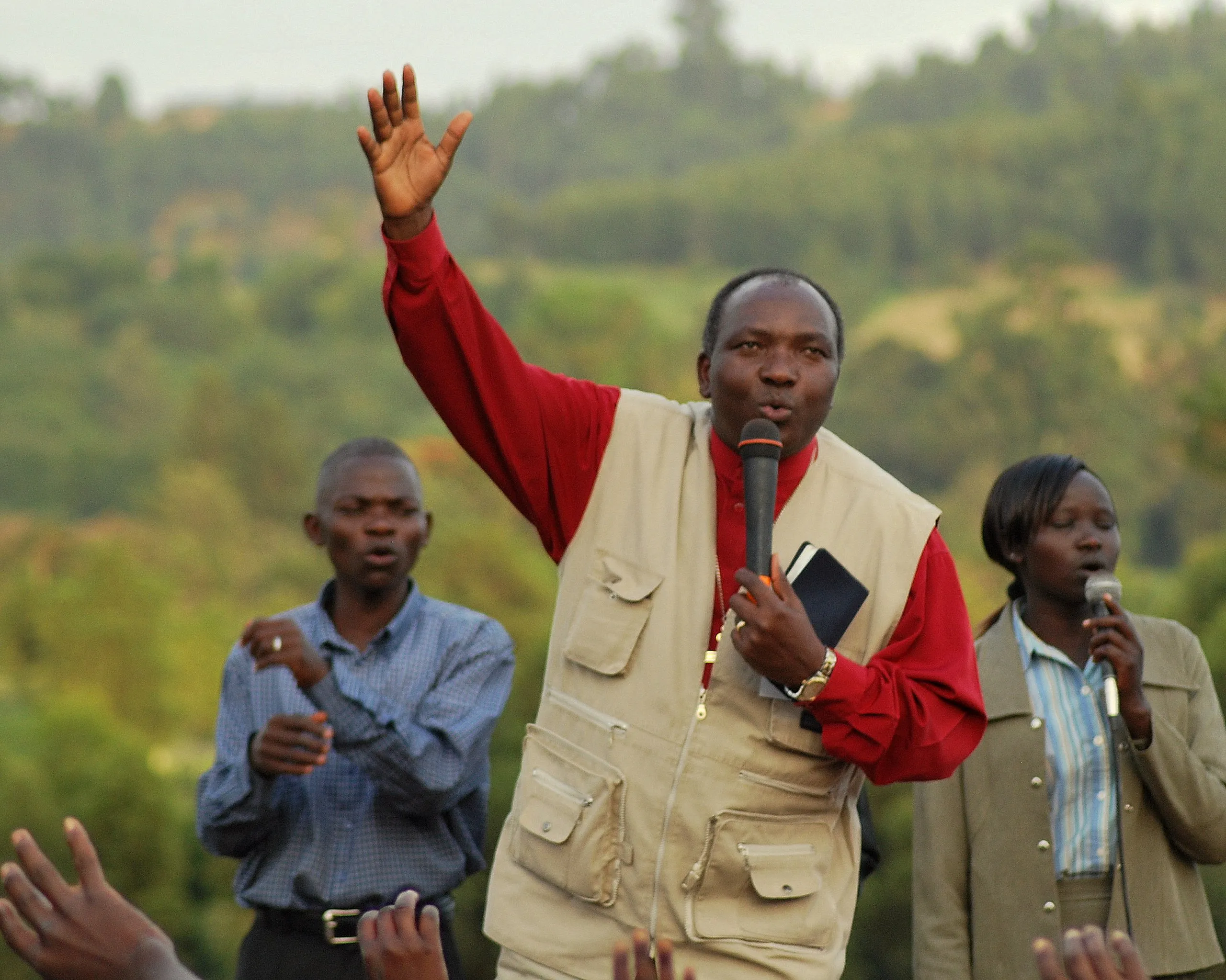 Man holding a microphone and a book, addressing an audience outdoors with two people standing behind him also holding microphones.