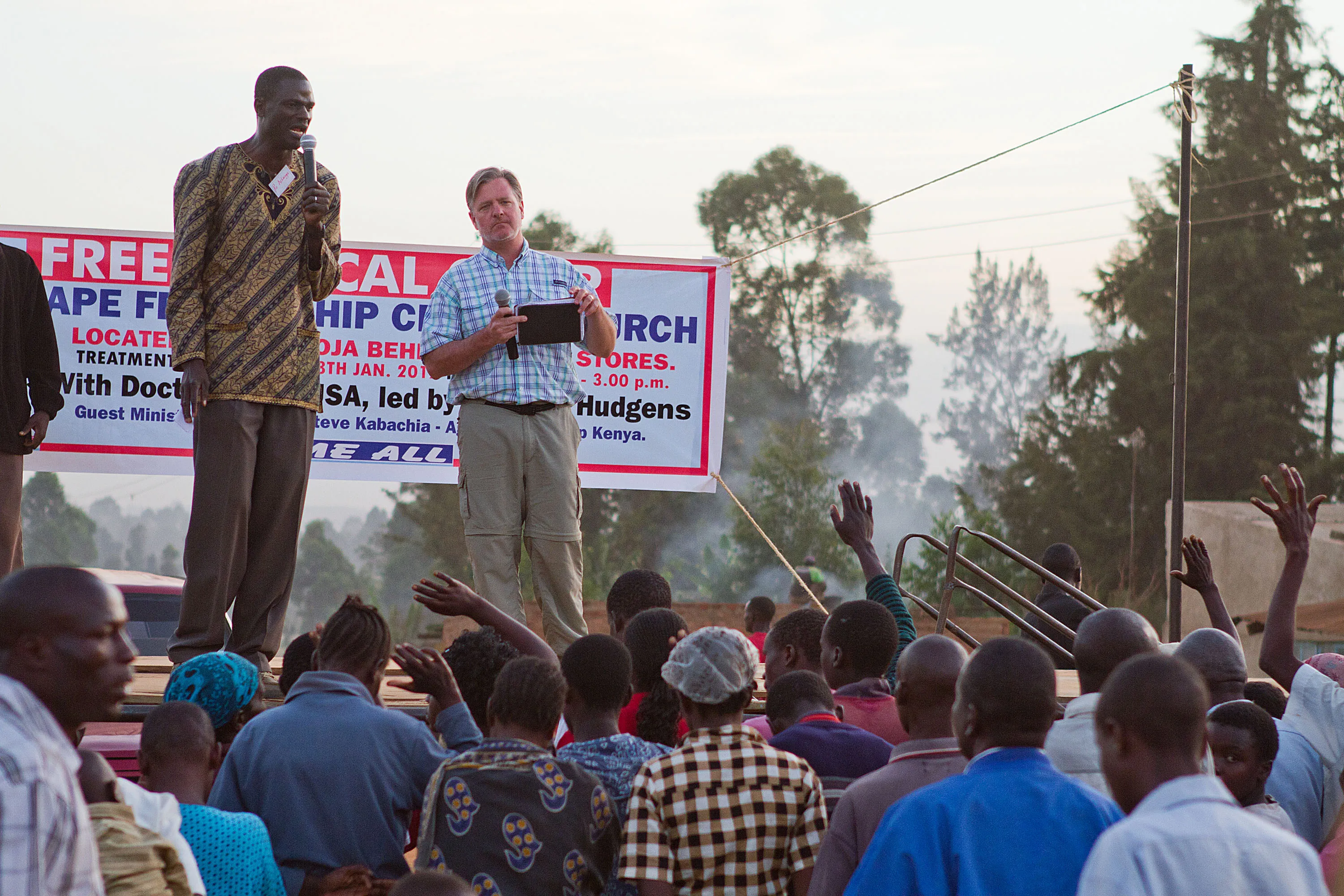 Two men speaking with microphones on a raised platform in front of a large crowd outdoors with trees in the background.