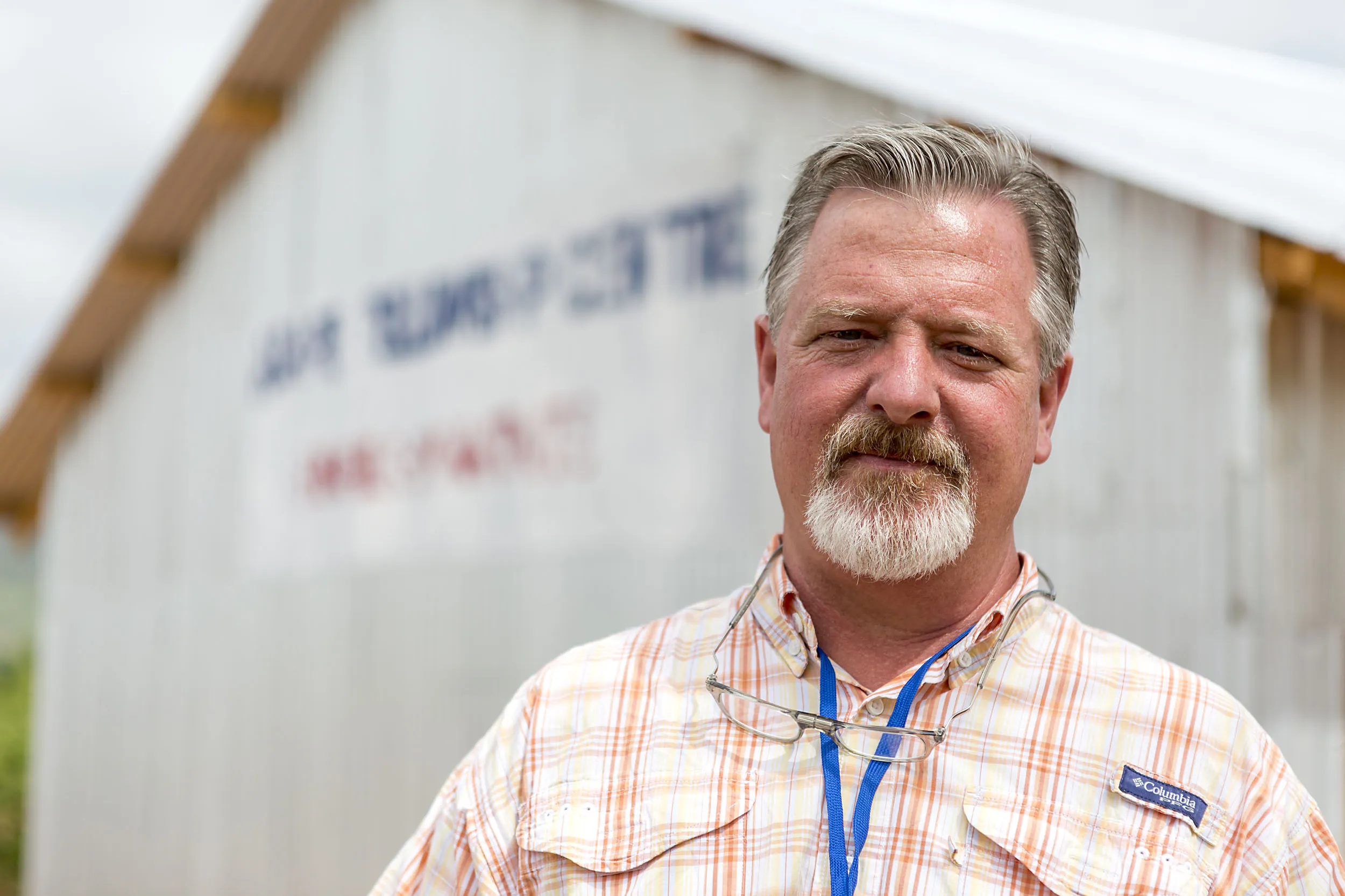 Middle-aged man with gray beard and mustache wearing a plaid shirt and glasses around his neck, standing outdoors in front of a white building.