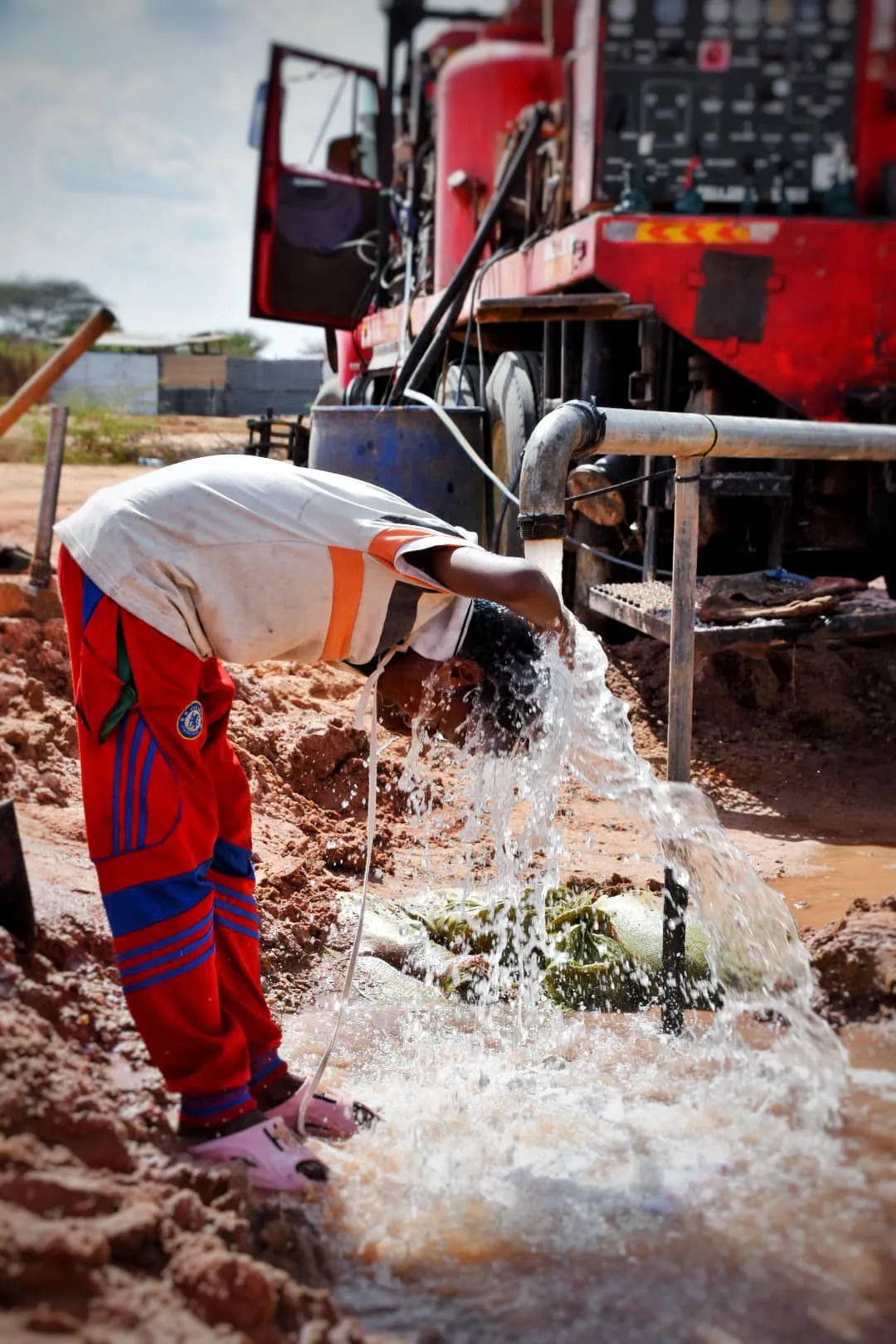 Child bending over to wash their face with water flowing from a metal pipe at a muddy construction site with a red machine in the background.