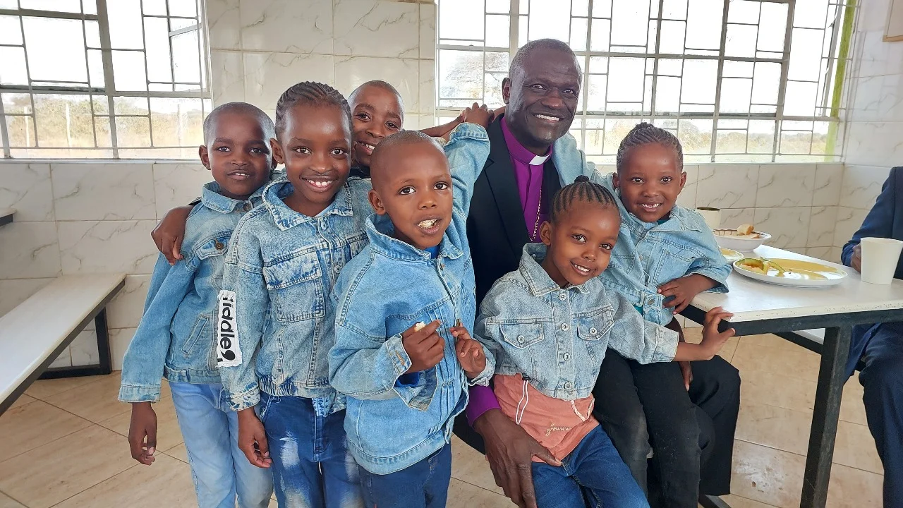 Group of smiling children wearing denim jackets gathered around a smiling man in a black suit with a purple clerical collar inside a room with tiled walls and windows.