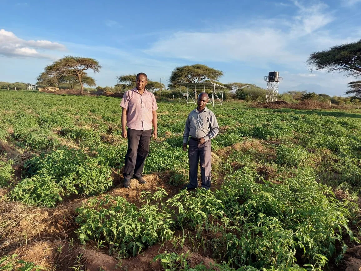 Two men standing in a green agricultural field with scattered trees and a water tank structure in the background under a partly cloudy sky.