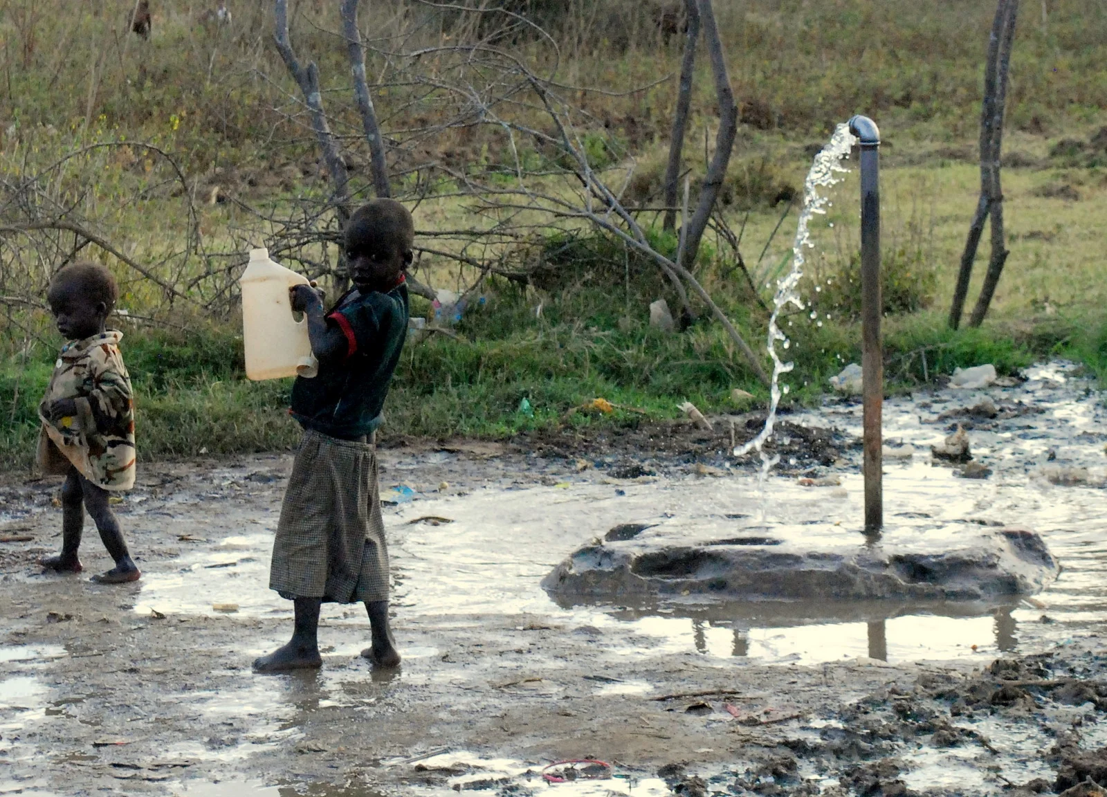 Two young children near a water pump with water flowing and muddy ground in a rural area.