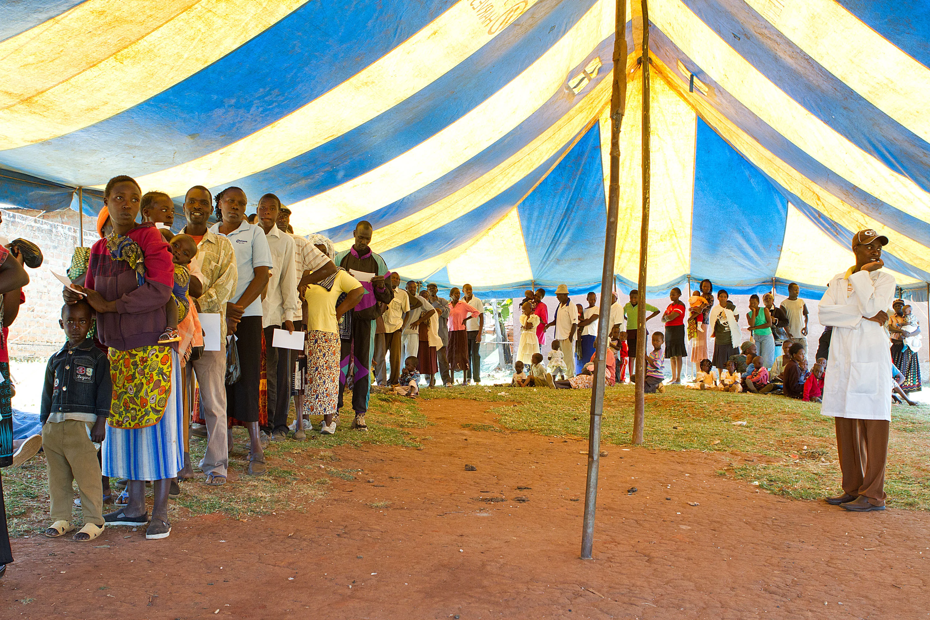 People standing in a long line under a large blue and yellow striped tent, with a man in a white coat observing nearby.