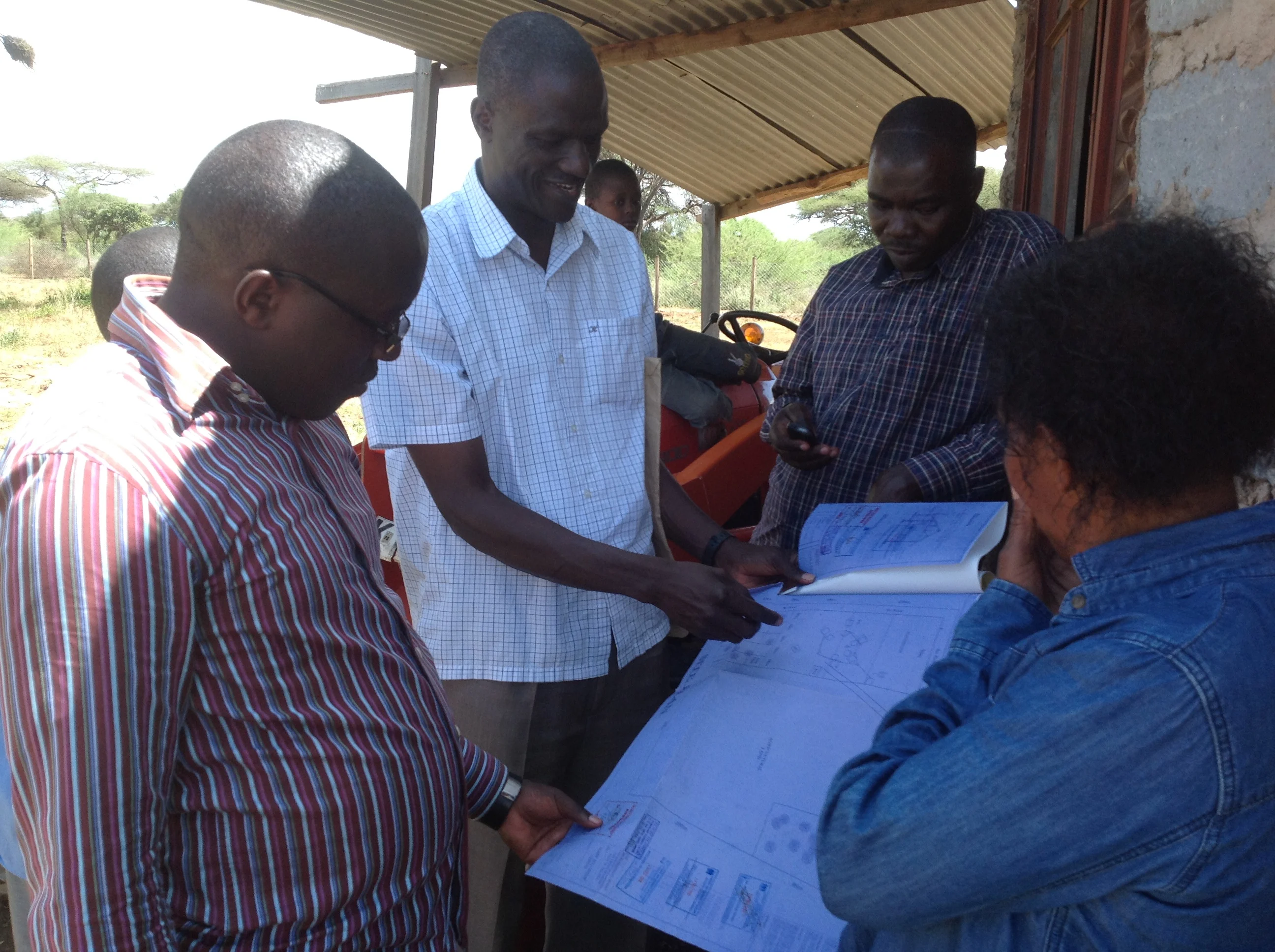 Four people discussing and examining large technical blueprints outside near a building and tractor.
