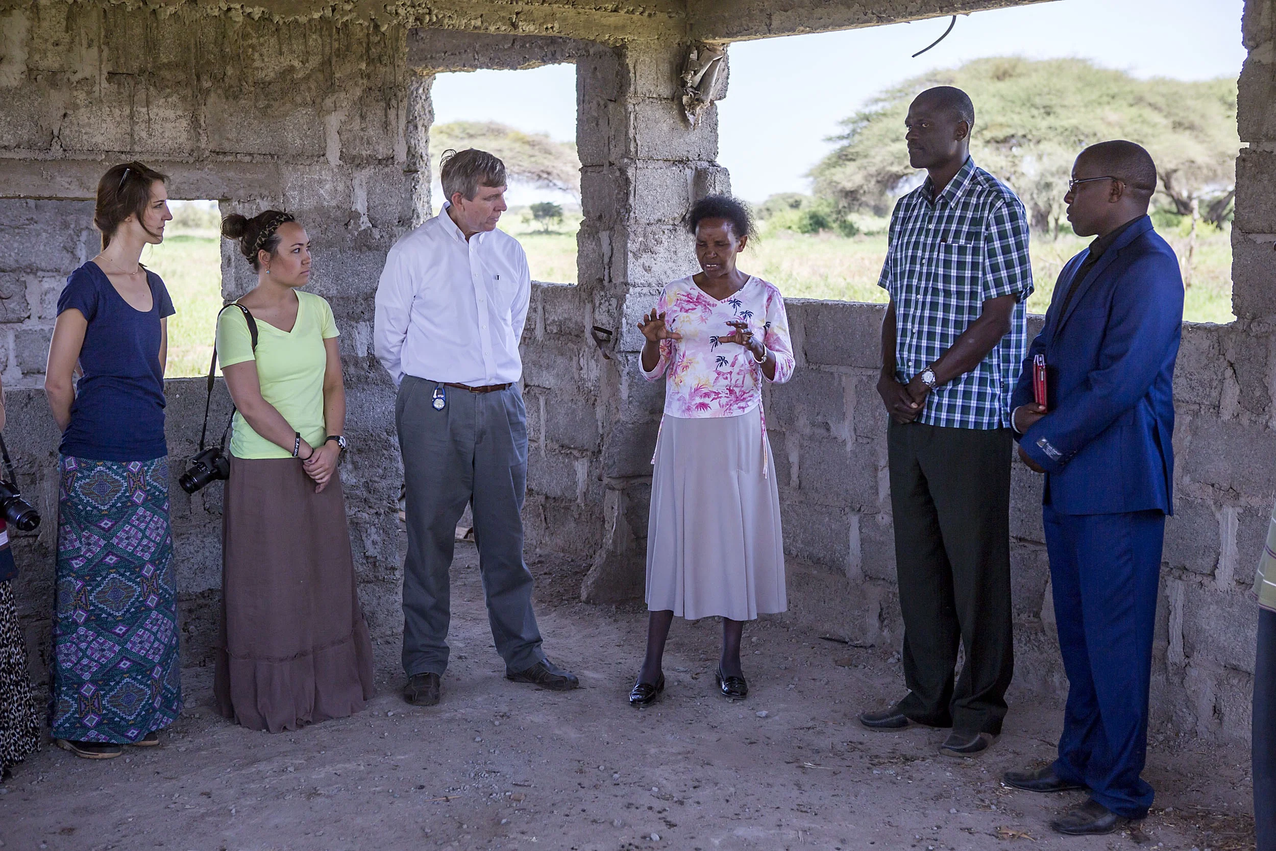A group of six people standing and listening to a woman speaking inside an unfinished building with concrete walls and window openings.