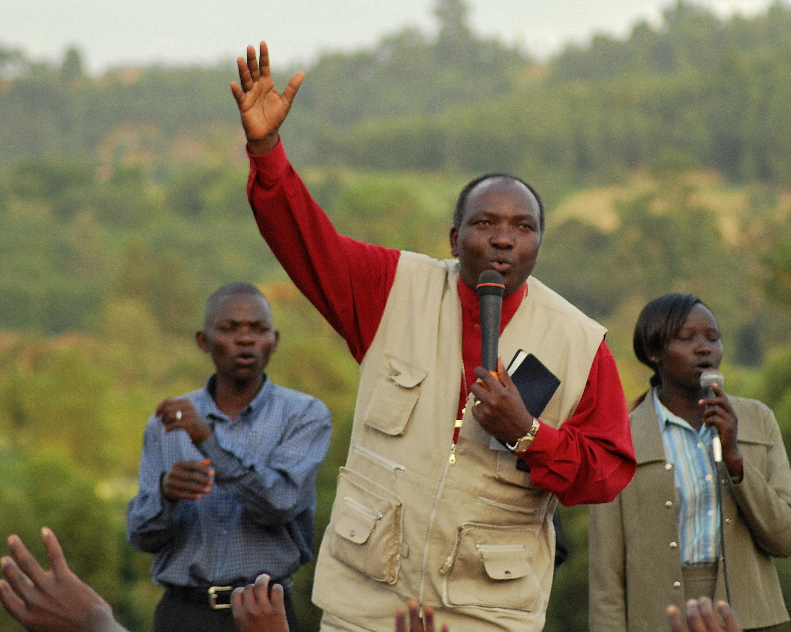 Man speaking into a microphone and raising his hand during an outdoor gathering with two people singing beside him.