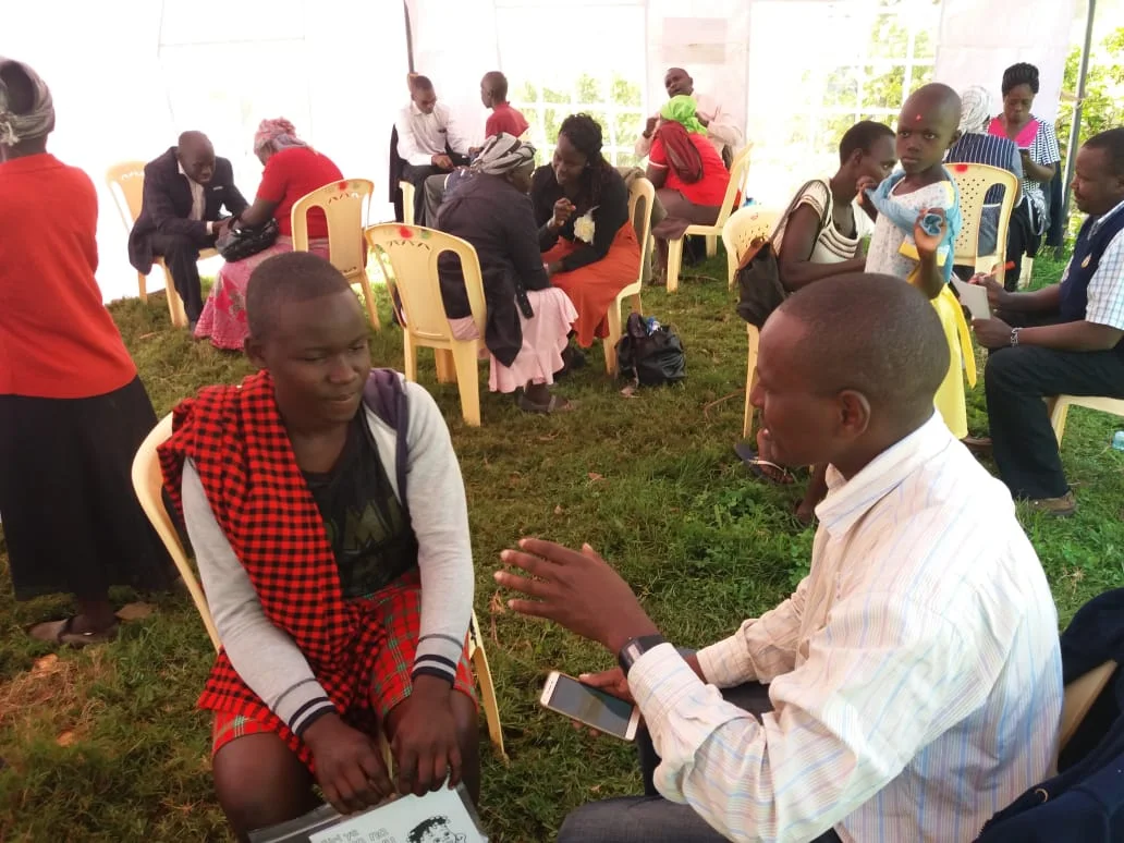 Several people engaged in seated conversations inside a tent on grass, with one man holding a smartphone and others in pairs talking.