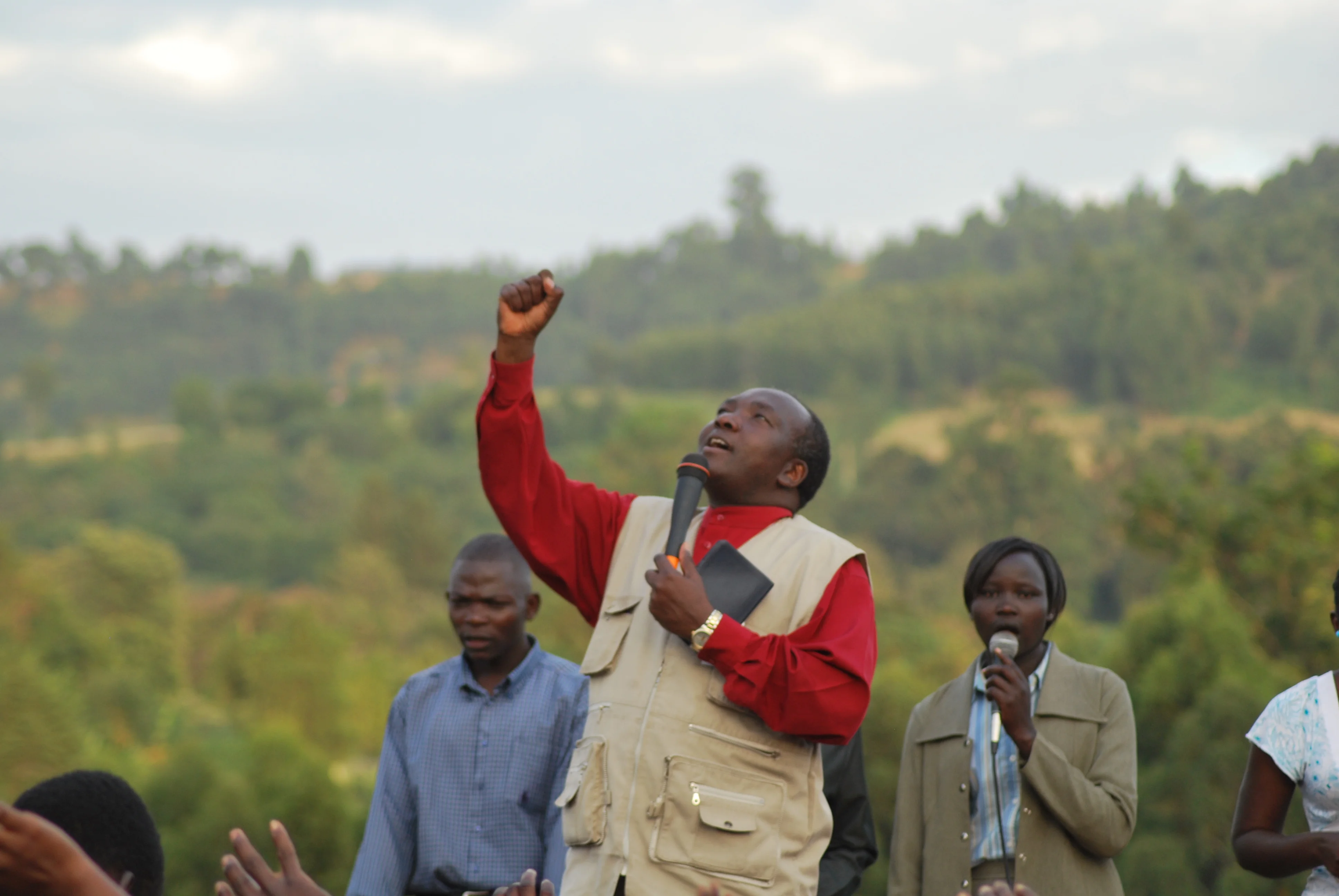 Man in a beige vest and red shirt holding a microphone and raising his fist, with another woman speaking into a microphone and people in the background outdoors.