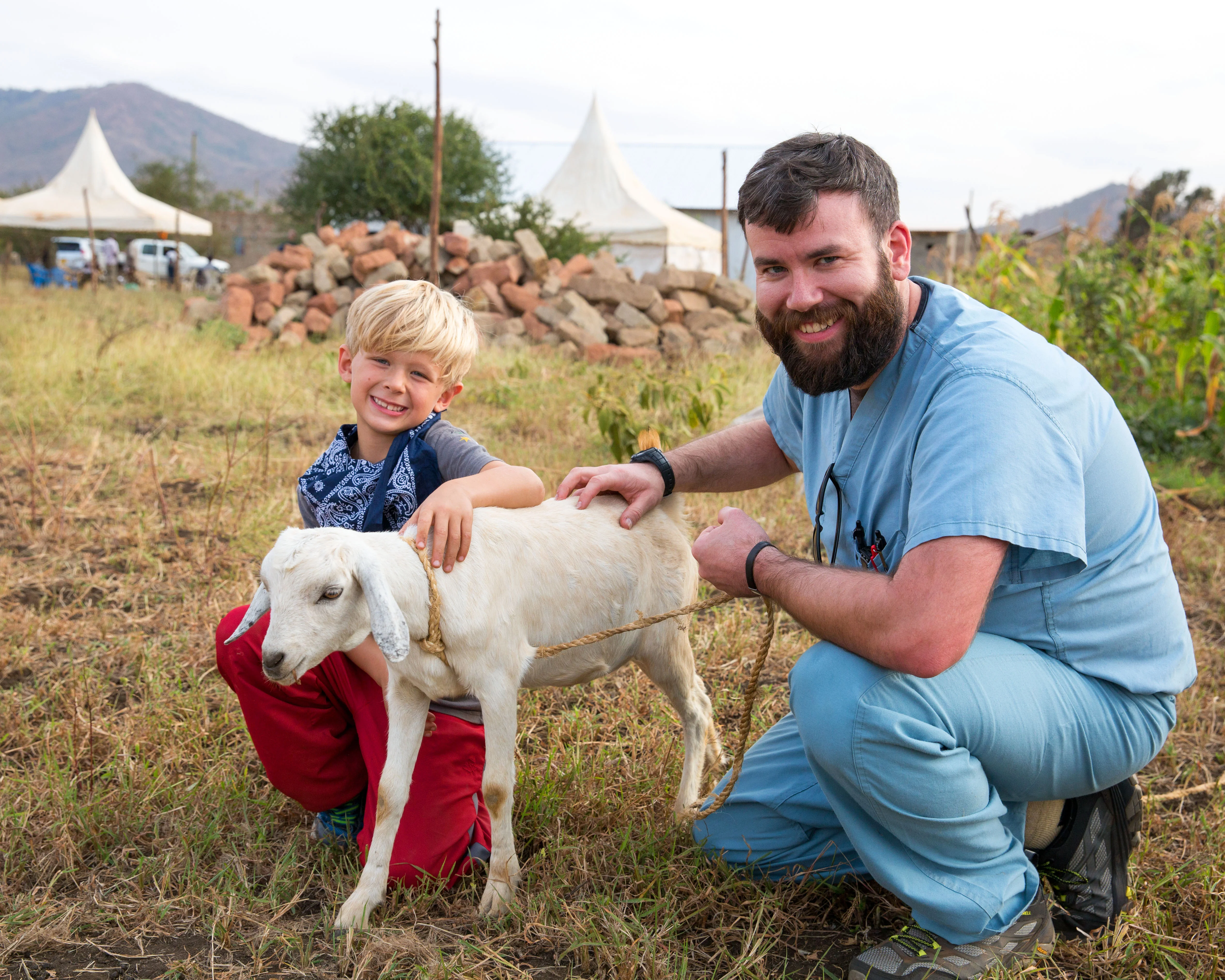 Smiling man in scrubs and young boy crouch beside a white goat in a grassy field with tents and mountains in the background.