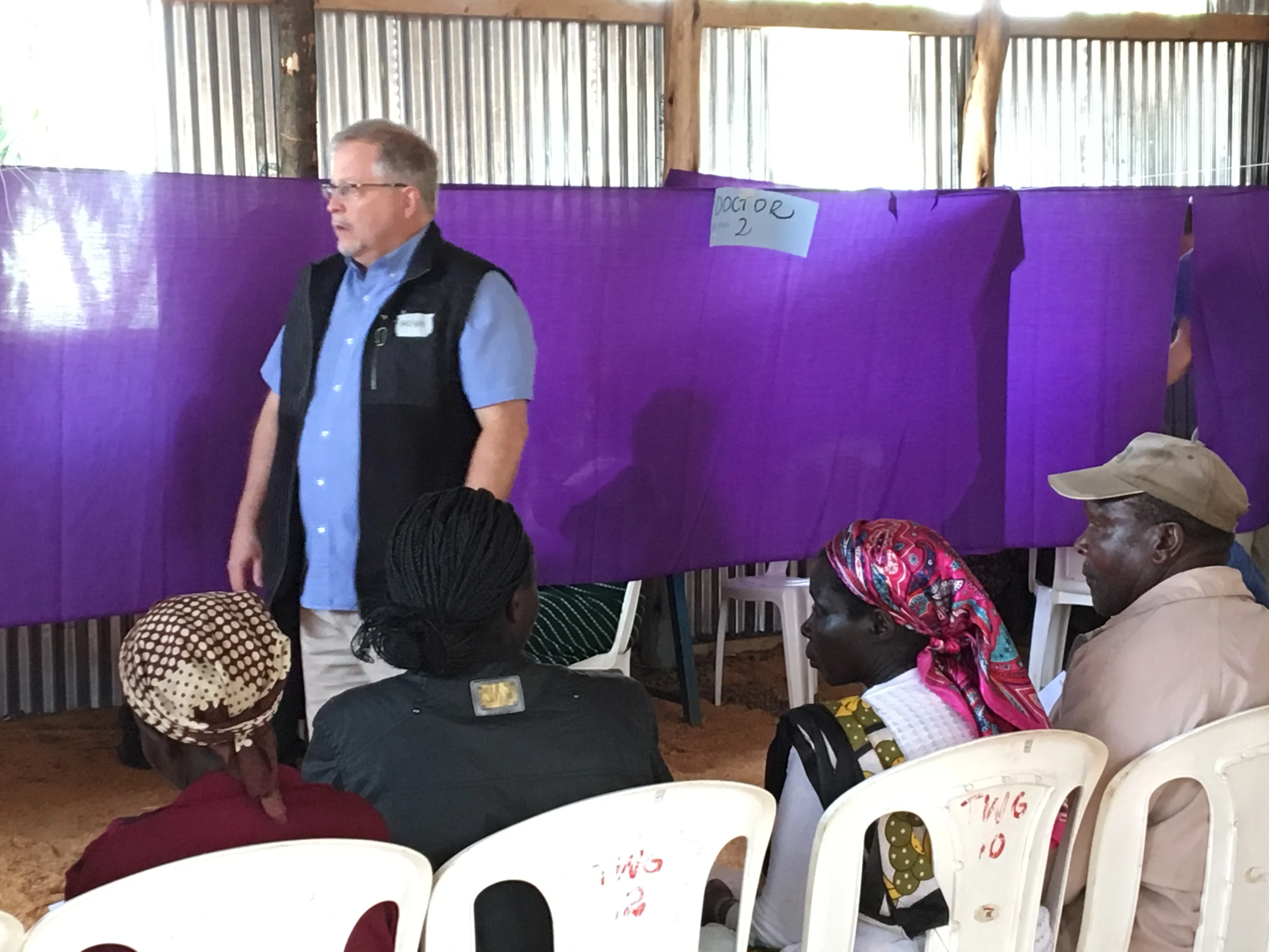 A man in a blue shirt and black vest stands speaking in front of a purple partition with a sign reading 'Doctor 2', while four people with varied head coverings sit facing him.