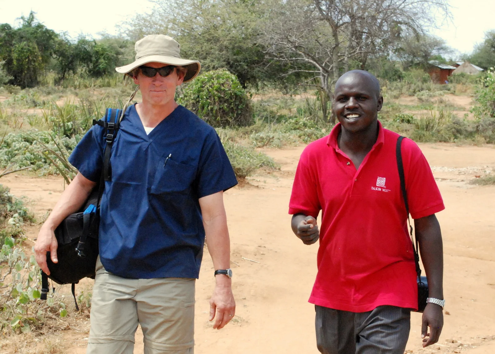 Two men walking on a dirt path in a rural area, one wearing a wide-brimmed hat and navy scrub top, the other in a red polo shirt smiling.