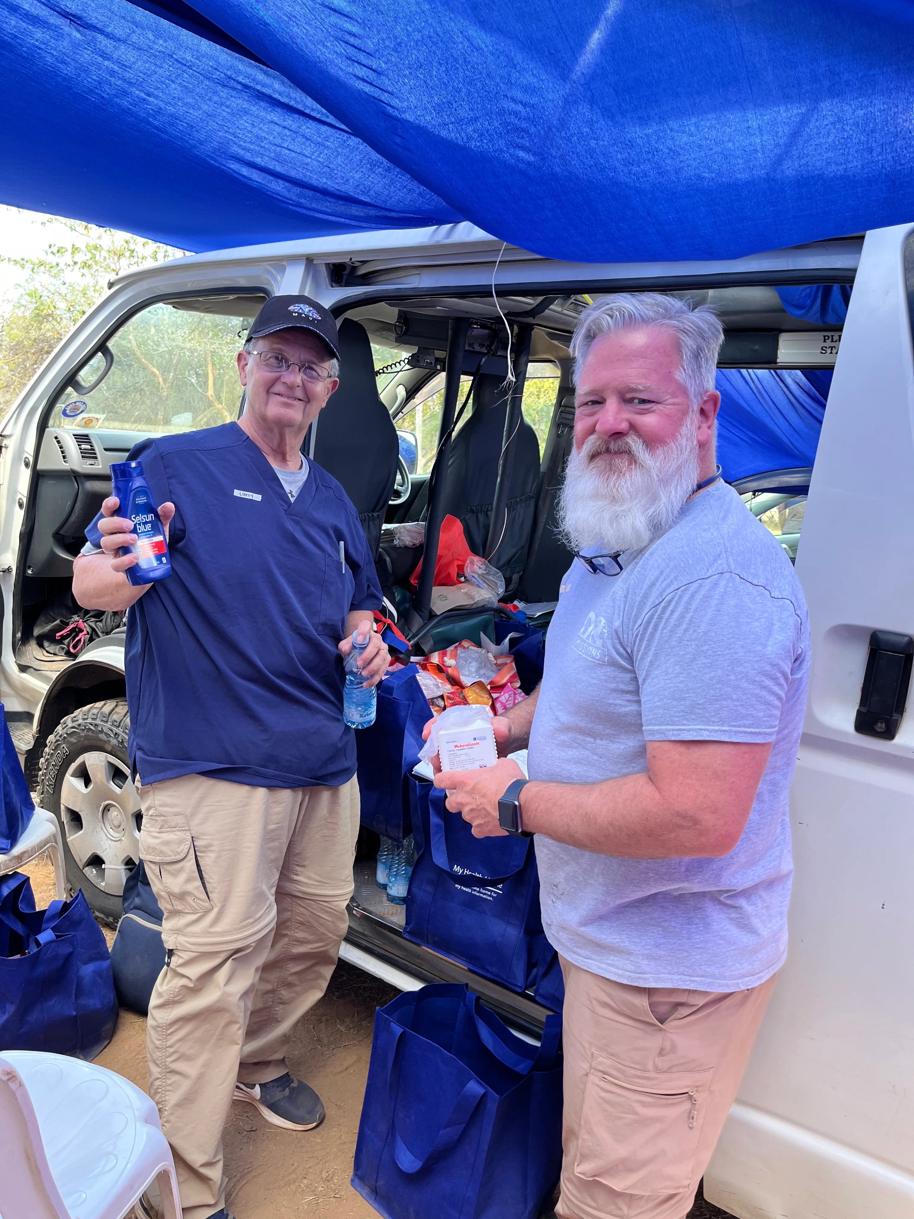Two men standing by an open white van, holding hygiene supplies with blue bags filled with items on the ground and in the van.