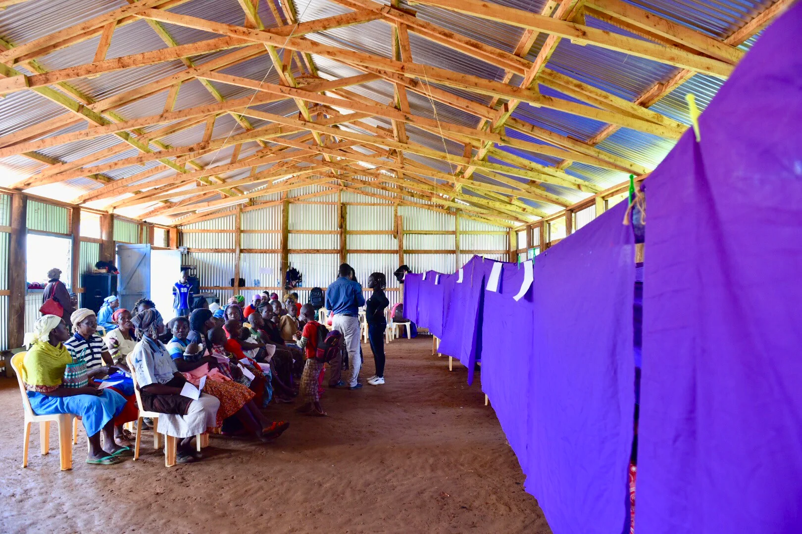 Group of people seated and standing inside a wooden and metal-roofed building with purple privacy curtains on one side.