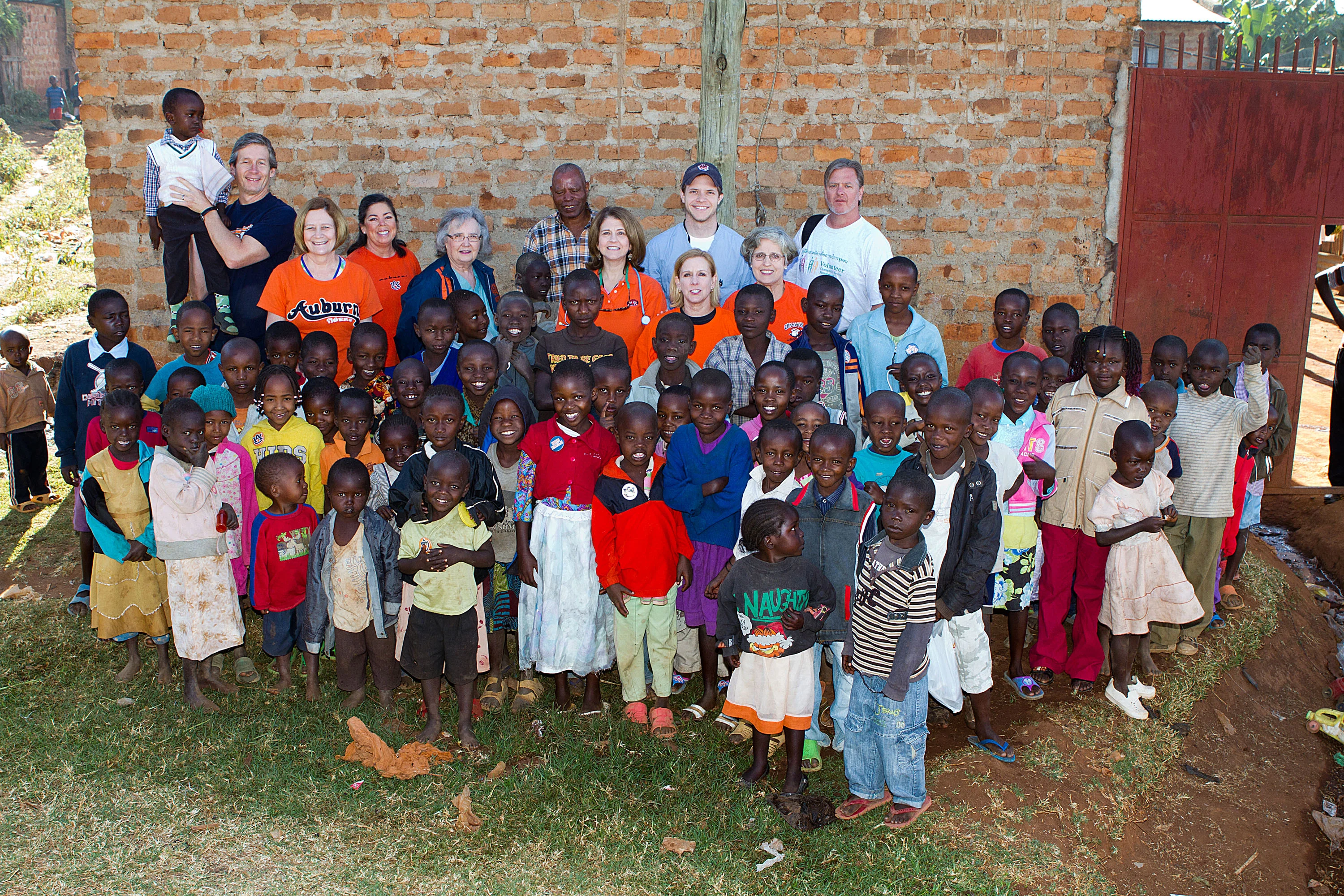 Group of smiling African children and several adults standing in front of a brick wall outside.