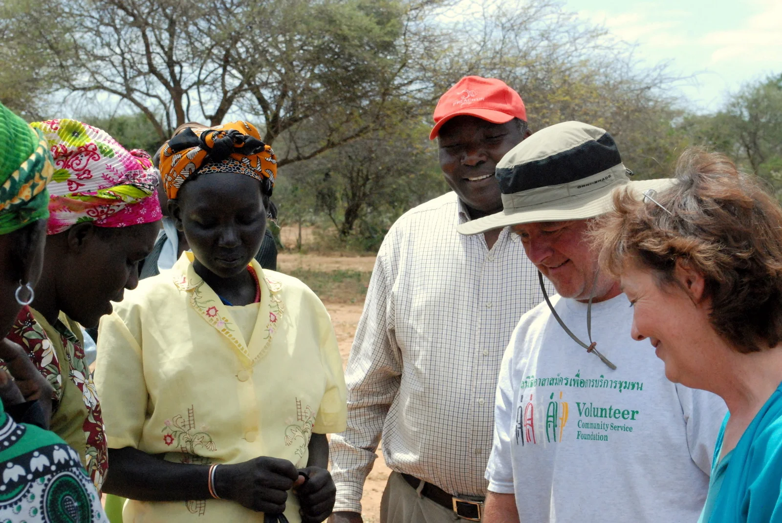 Group of diverse adults outdoors, including two women in colorful headscarves, a man in a red cap, and two volunteers engaged in conversation.