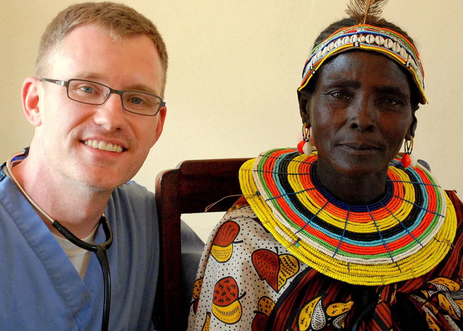 A smiling male doctor wearing glasses and a stethoscope poses beside an older woman dressed in colorful traditional beaded jewelry and clothing.