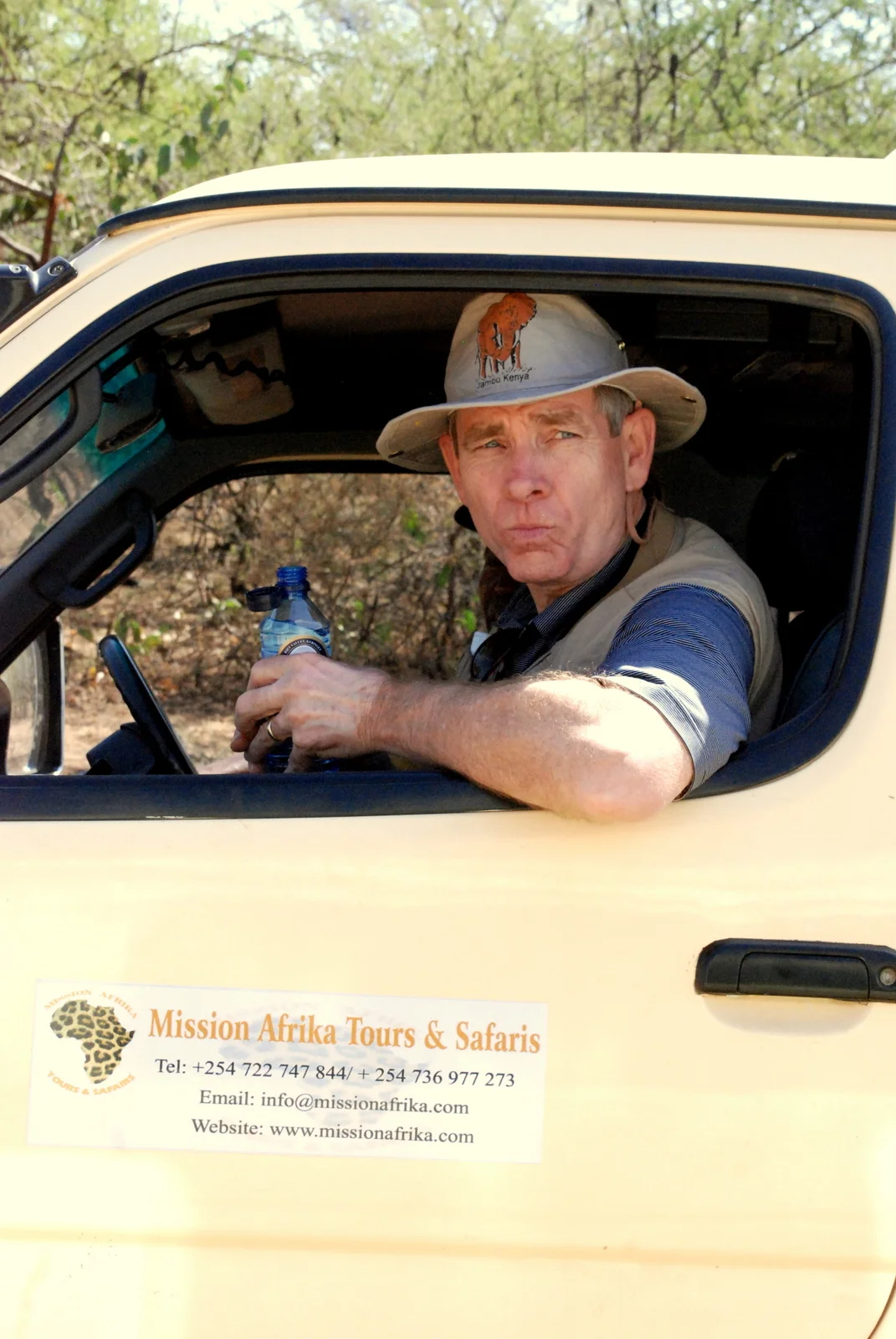 Man wearing a safari hat sitting inside a safari vehicle holding a water bottle, with a Mission Afrika Tours & Safaris sign on the vehicle door.